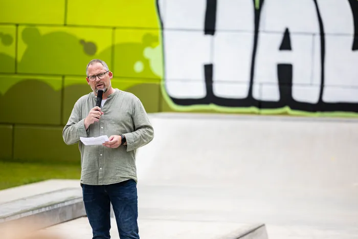 Ein Mann mit Bart hält ein Mikrofon in der Hand und spricht. Im Hintergrund ist ein Skatepark mit Graffiti zu sehen.