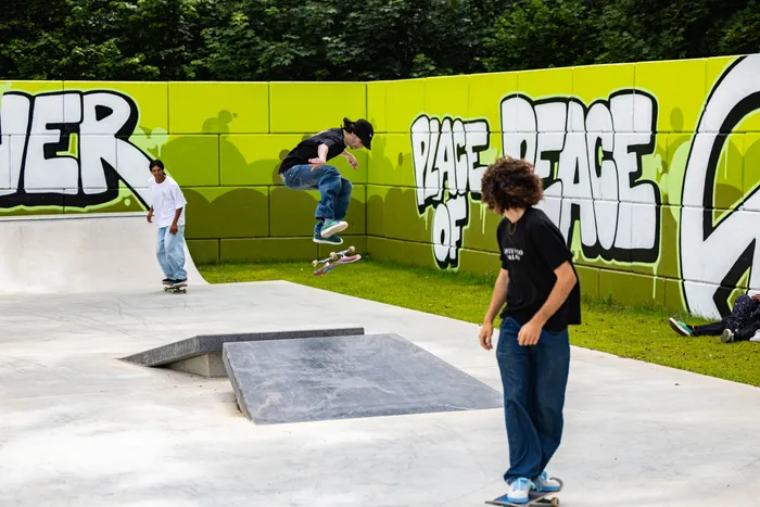 Drei junge Menschen stehen und fahren auf Skateboards in einem neu eröffneten Skatepark. Im Hintergrund eine grüne Wand mit Graffiti-Schriftzügen.
