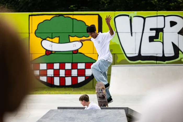 Ein junger Mann fährt auf einem Skateboard über eine Rampe in einem Skatepark. Im Hintergrund ist eine Wand mit Graffiti und einer großen Burger-Darstellung zu sehen, während ein weiterer Junge zuschaut.