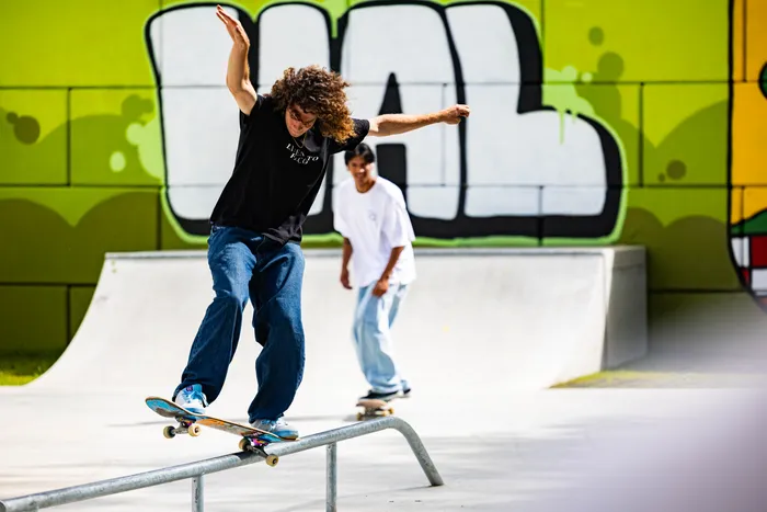 Ein junger Skater mit lockigem Haar fährt auf einem Skatepark-Geländer, während ein zweiter Skater im Hintergrund auf einem Rampenabschnitt fährt. Im Hintergrund ist eine grüne Wand mit Graffiti zu sehen.