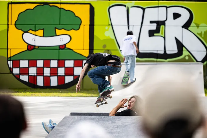 Ein Skater springt mit seinem Board in der Luft, während ein Mann im Hintergrund überrascht reagiert. Im Hintergrund ist ein Graffiti mit einem Burger und der Aufschrift „WER“ zu sehen.