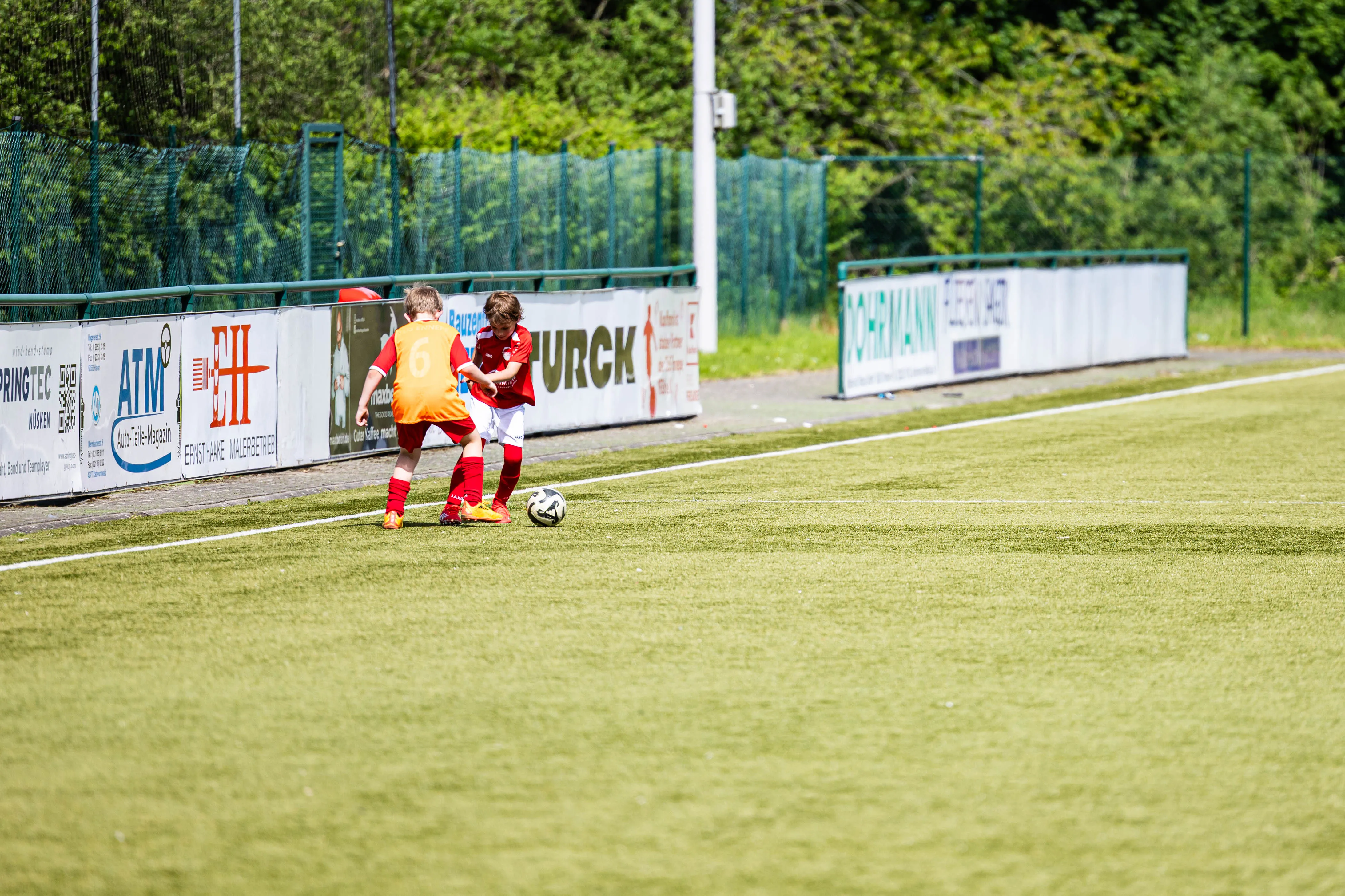 Zwei junge Fußballspieler in orange und rot spielen auf einem grünen Rasenfeld, umgeben von Werbebannern und einem Zaun.