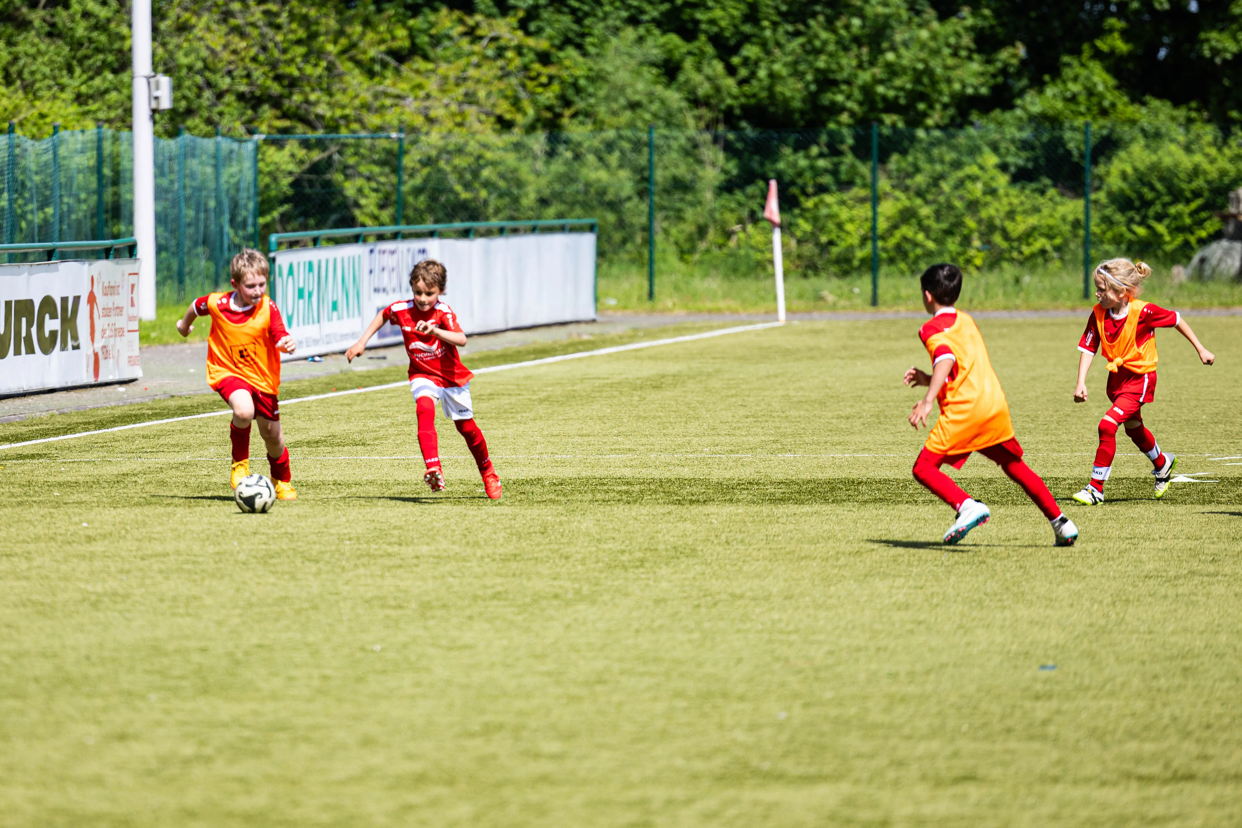 Vier Kinder in Fußballtrikots spielen auf einem grünen Rasenfeld. Zwei tragen orangefarbene Westen, die anderen rote Trikots.
