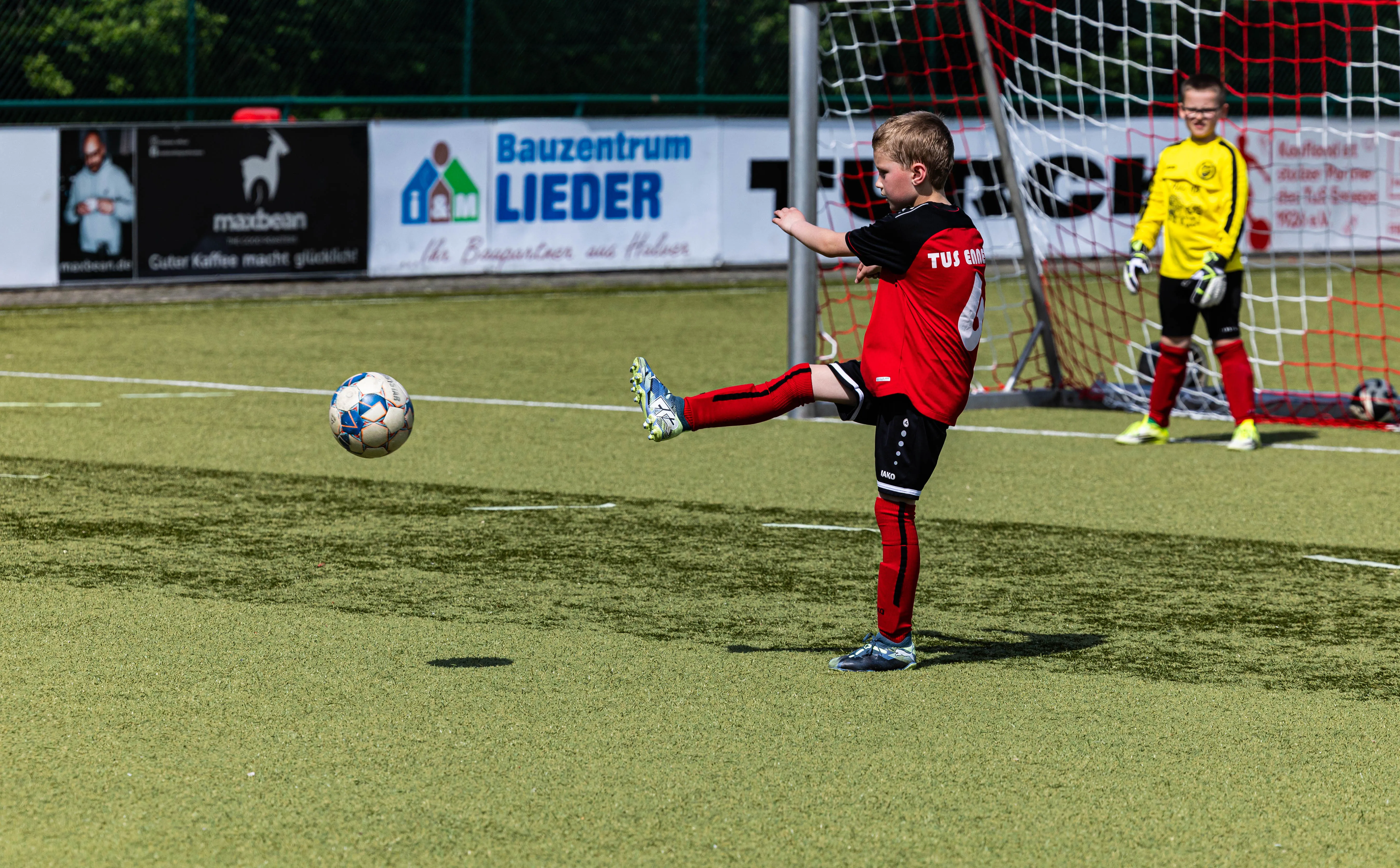 Ein Jugendfußballspieler in rotem Trikot schießt auf das Tor, während der Torwart in gelber Kleidung bereit steht.