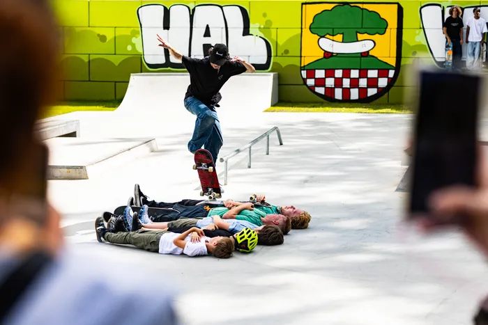 Ein Skateboarder springt in der Luft über eine Gruppe von Jungen, die auf dem Boden liegen und zusehen. Im Hintergrund ist eine grüne Wand mit Graffiti und Skatepark-Elemente zu sehen.