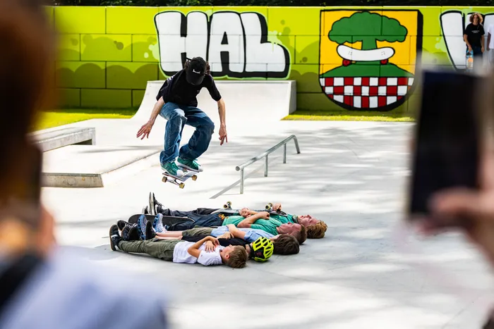 Ein Skater springt mit seinem Board über eine Gruppe von Kindern, die auf dem Boden liegen. Im Hintergrund ist eine bunte Wand mit Graffiti und Skatepark-Elemente zu sehen.