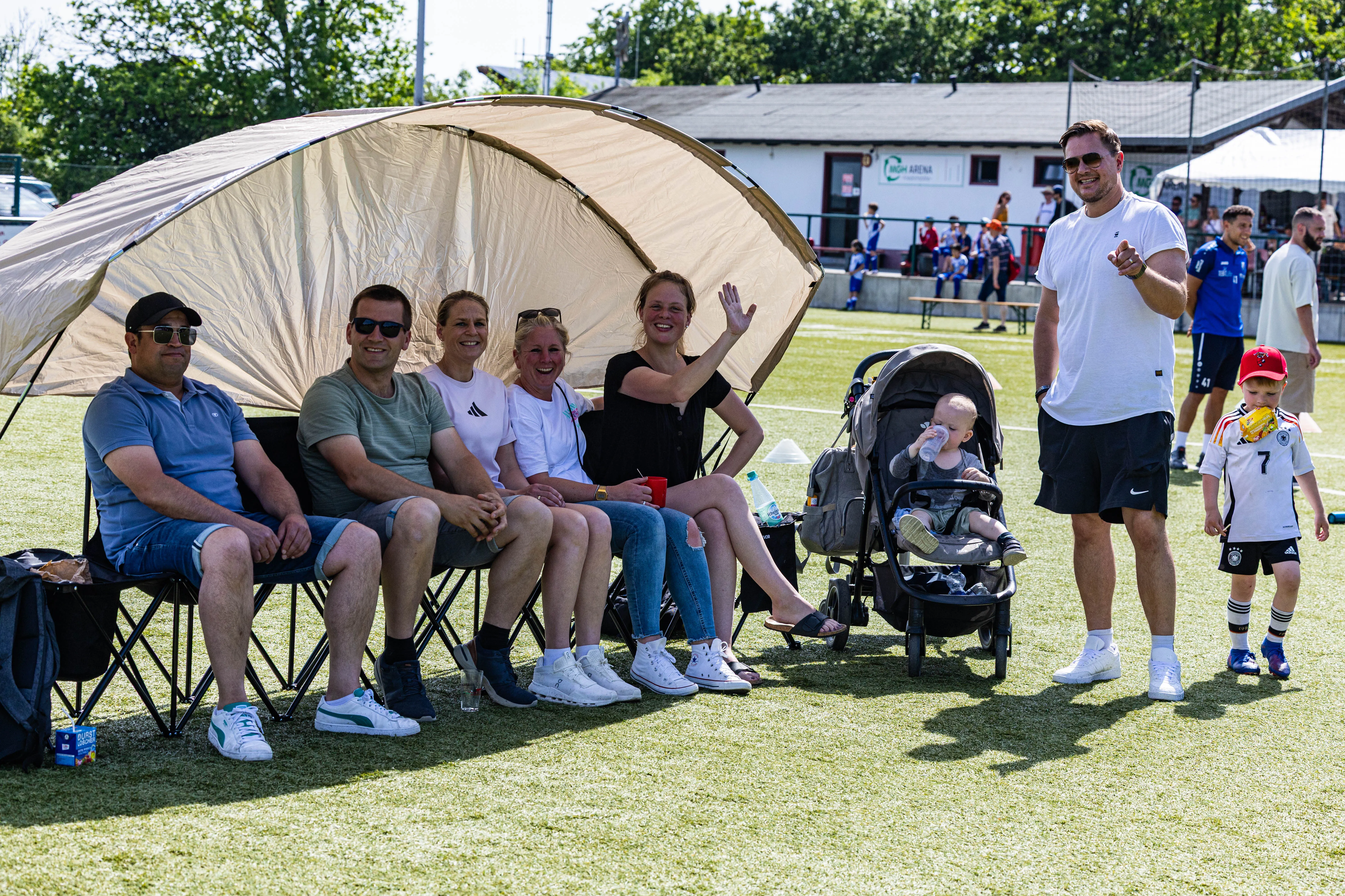 Zuschauer sitzen und stehen am Spielfeldrand eines Fußballturniers, einige unter einem Sonnensegel. Ein Mann winkt in die Kamera, während ein Baby im Kinderwagen sitzt.