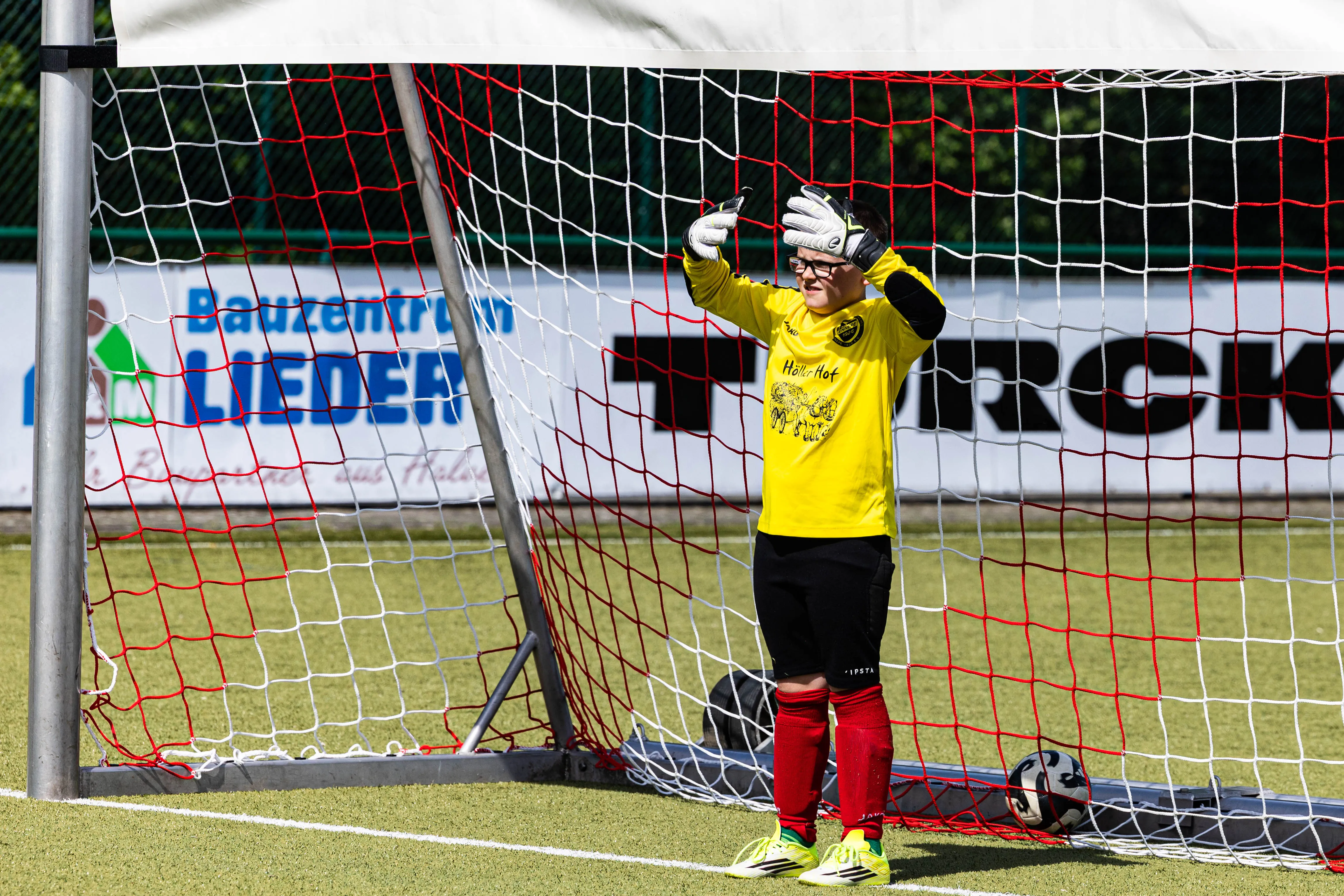 Torwart in gelbem Trikot und roten Stutzen steht vor dem Tor und hält die Hände hoch. Im Hintergrund sind Werbebanner und Bäume zu sehen.