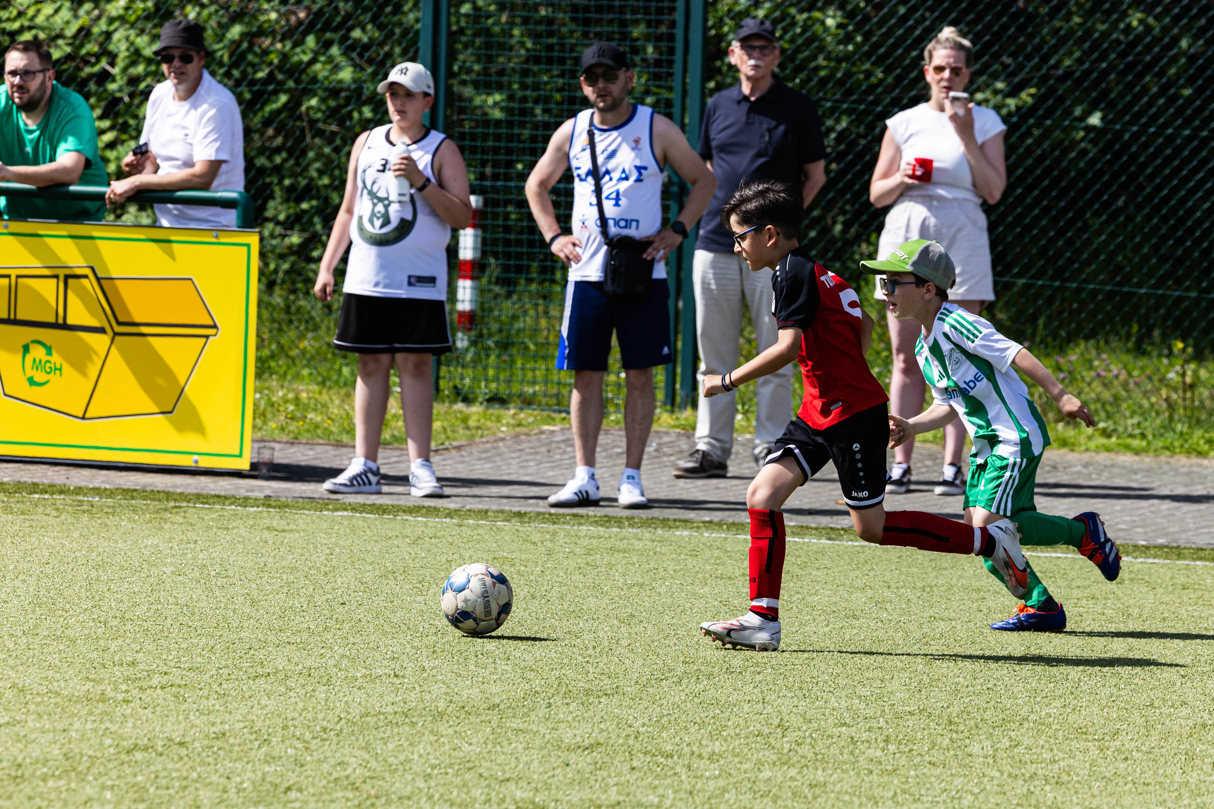 Zwei junge Fußballspieler in Aktion auf einem grünen Kunstrasenfeld, während Zuschauer im Hintergrund zusehen.