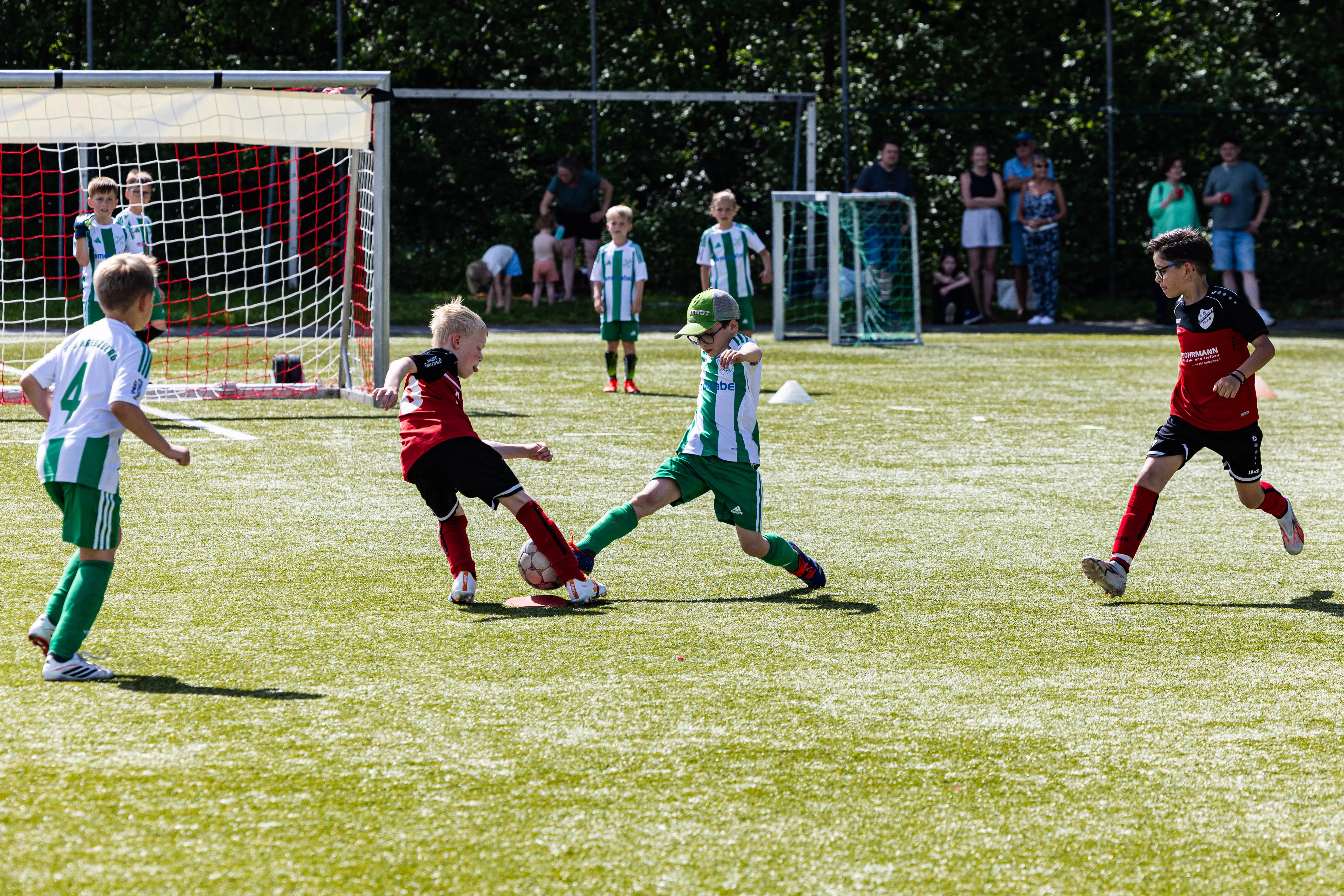 Zwei Jugendfußballspieler kämpfen um den Ball: Ein Spieler in Rot und Schwarz wird von einem Spieler in Grün angegriffen. Im Hintergrund weitere Spieler und Zuschauer am Spielfeldrand.