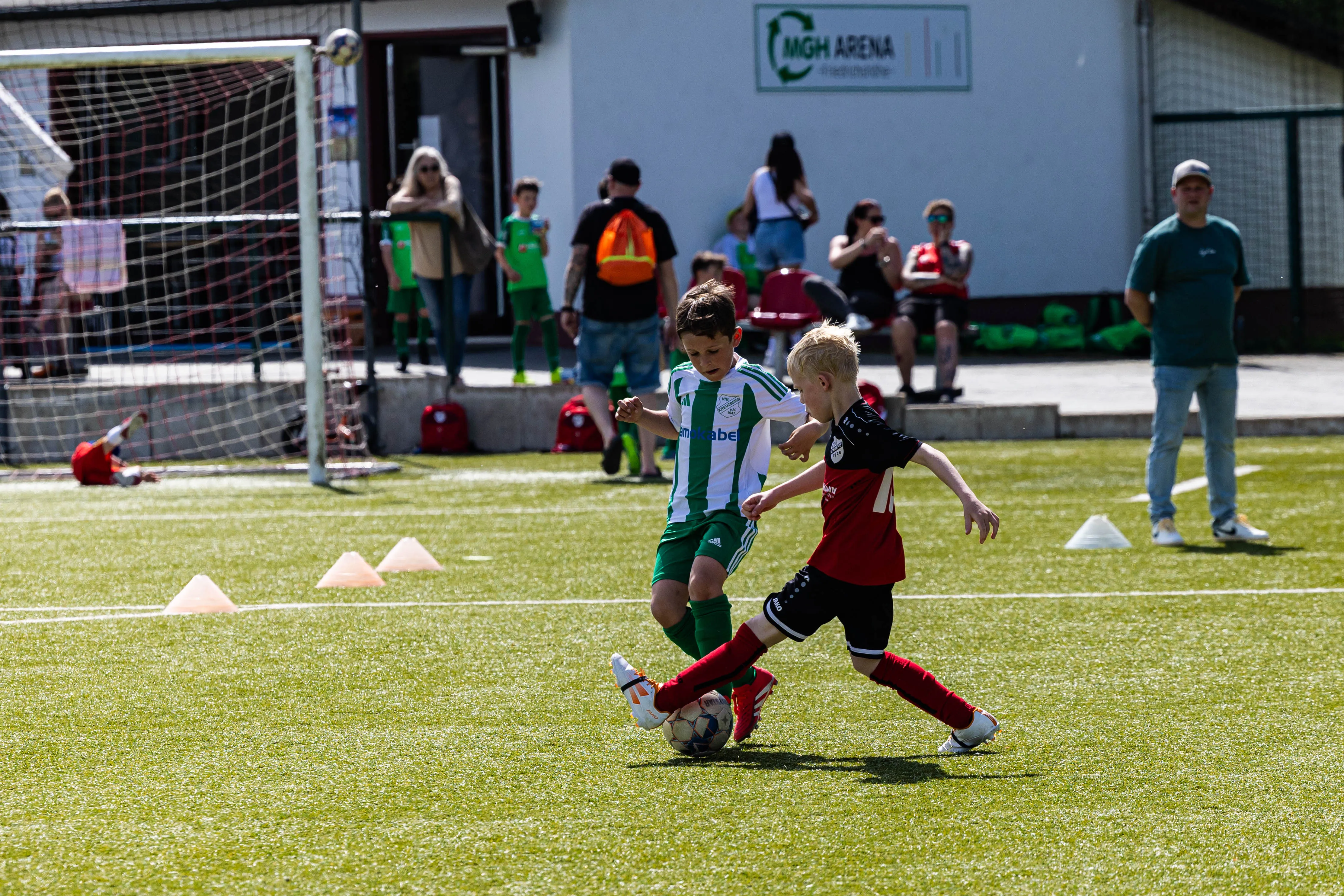 Zwei junge Fußballspieler in Vereinskleidung kämpfen um den Ball auf einem grünen Rasenfeld, während im Hintergrund Zuschauer zusehen.