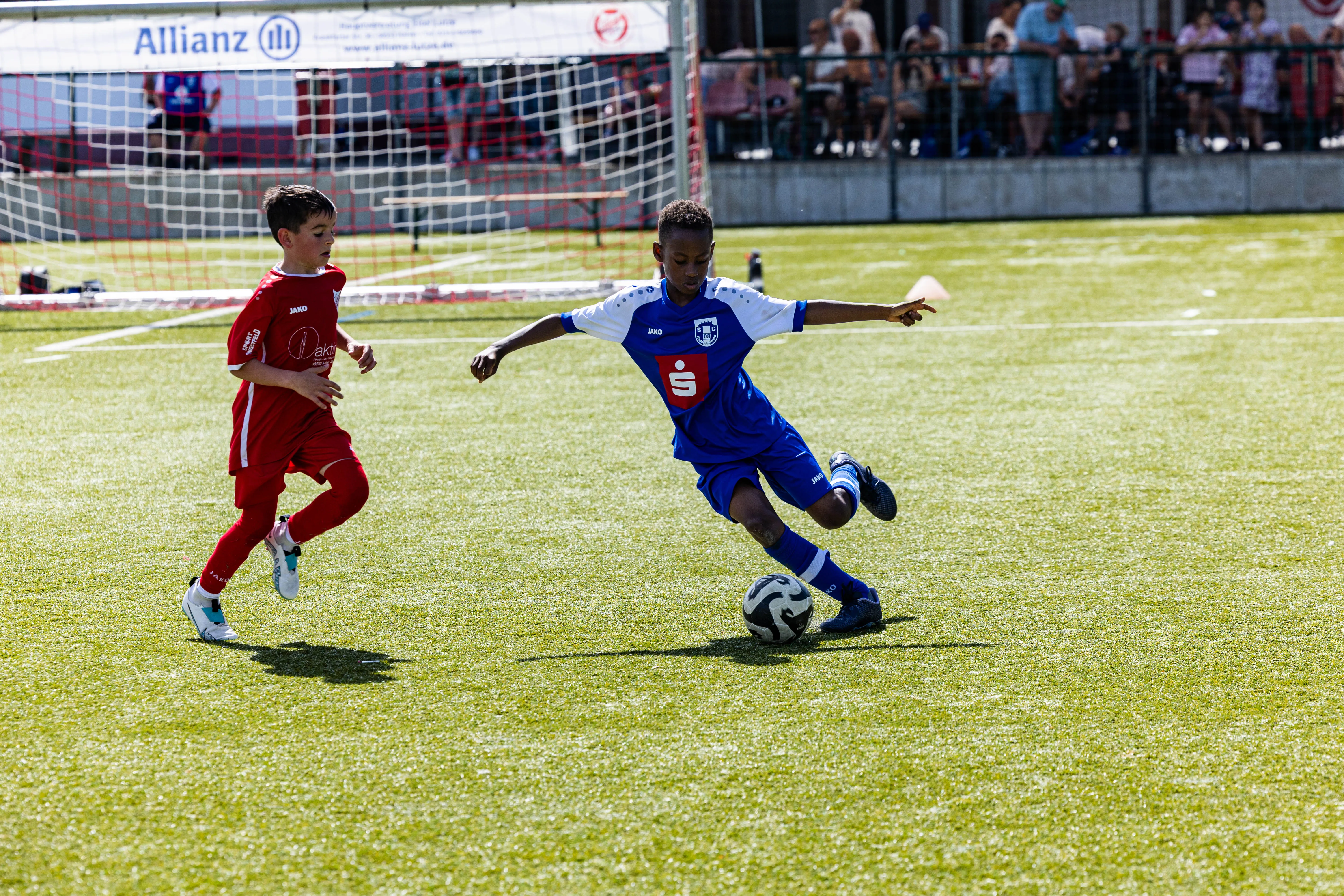 Zwei junge Fußballspieler im Wettkampf: Ein Spieler in rotem Trikot verfolgt einen anderen in blauem Trikot, der den Ball führt.