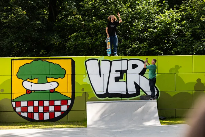 Eine Person steht auf einer Betonmauer vor einer bunten Wand mit Graffiti und winkt. Ein zweiter Mensch steht daneben und scheint etwas zu zeigen. Im Hintergrund ist dichter Grünbewuchs zu sehen.