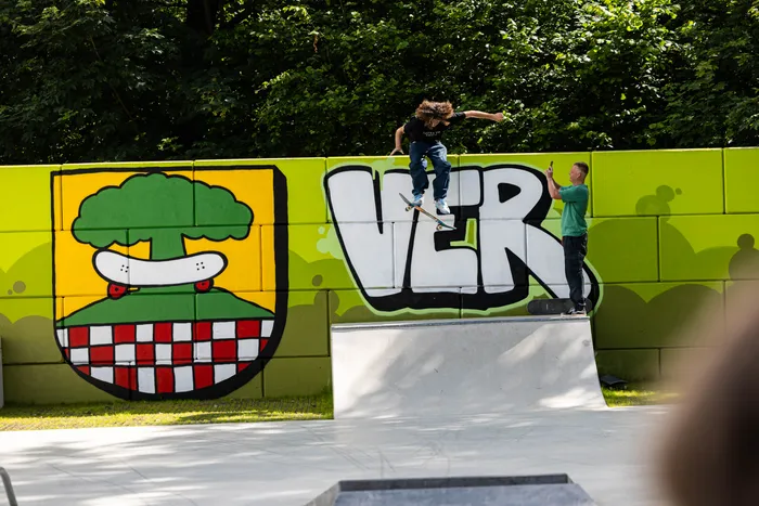 Ein Skater springt auf einem Skatepark vor einer Wand mit Graffiti und einem Wappen. Ein Zuschauer filmt mit dem Handy.