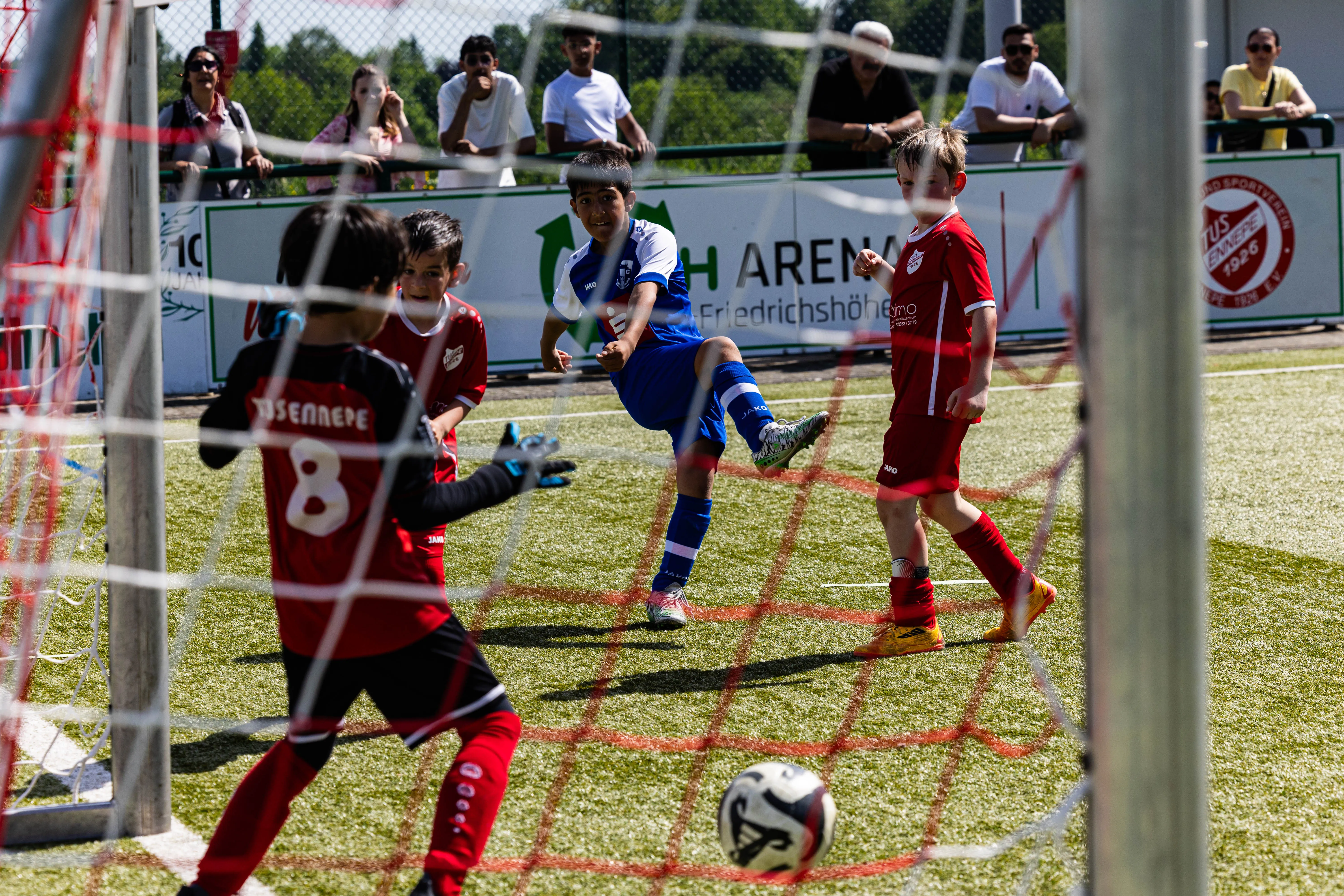 Ein Jugendlicher in blauem Trikot schießt im Fußballspiel gegen einen Torwart in rotem Trikot. Im Hintergrund stehen weitere Spieler und Zuschauer auf einer Tribüne.