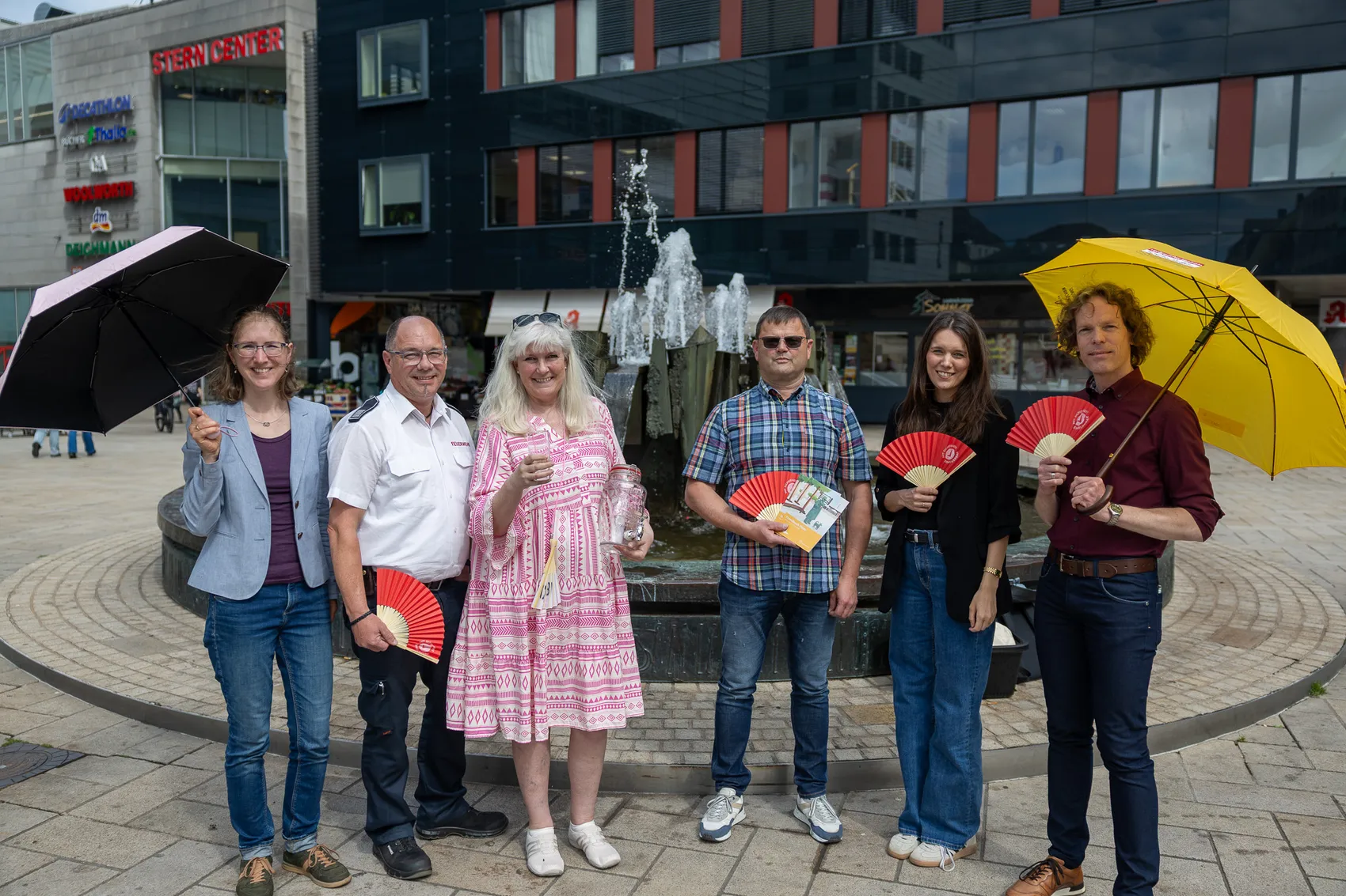 Eine Gruppe von sechs Personen steht vor einem Springbrunnen und hält Regenschirme und Fächer in den Händen. Im Hintergrund befindet sich ein modernes Gebäude mit der Aufschrift „Stern-Center“.