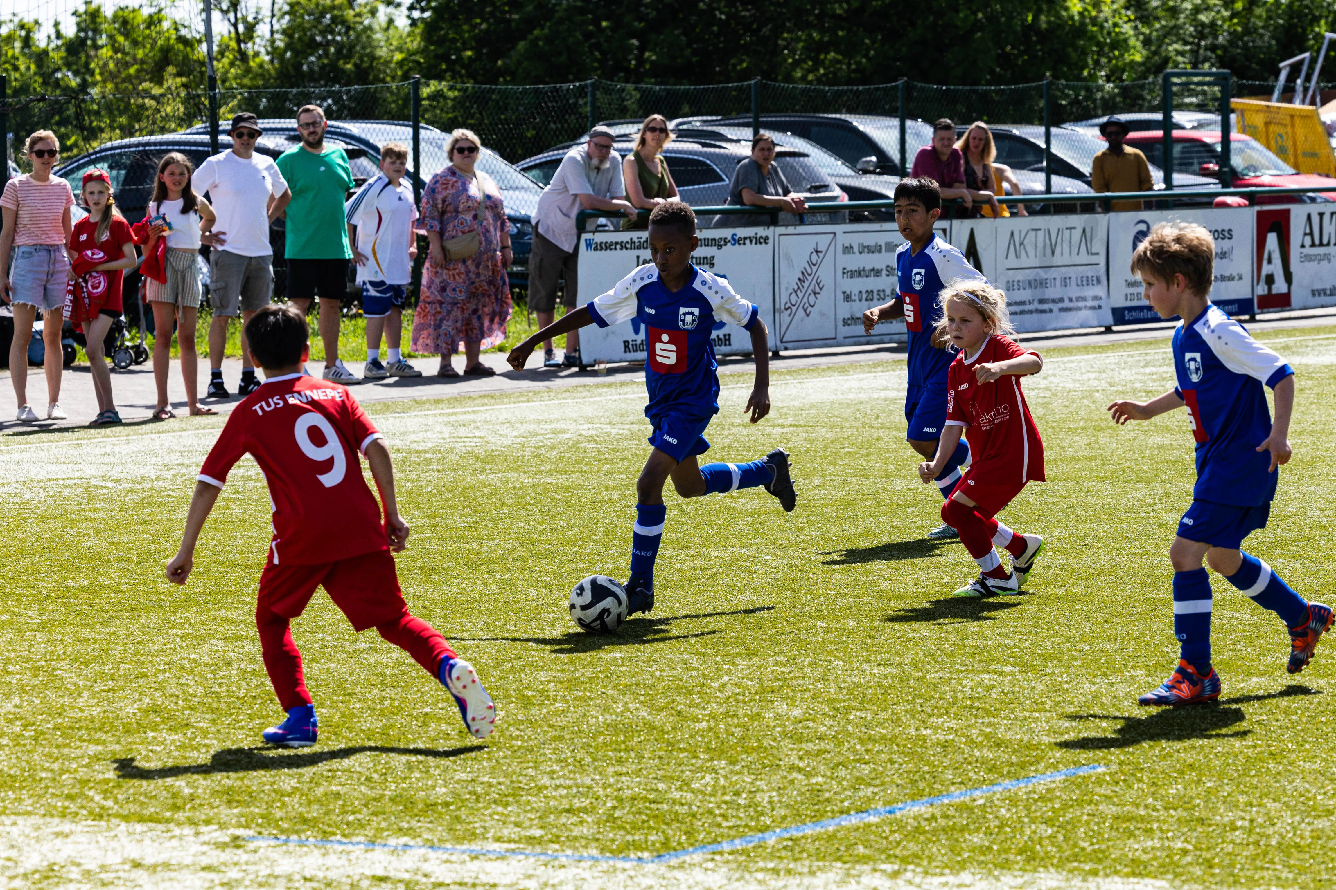 Jugendfußballspiel auf einem Rasenplatz: Zwei Spieler in roten Trikots verfolgen einen Spieler in blauem Trikot, der den Ball führt. Im Hintergrund stehen Zuschauer und sind Werbebanner zu sehen.