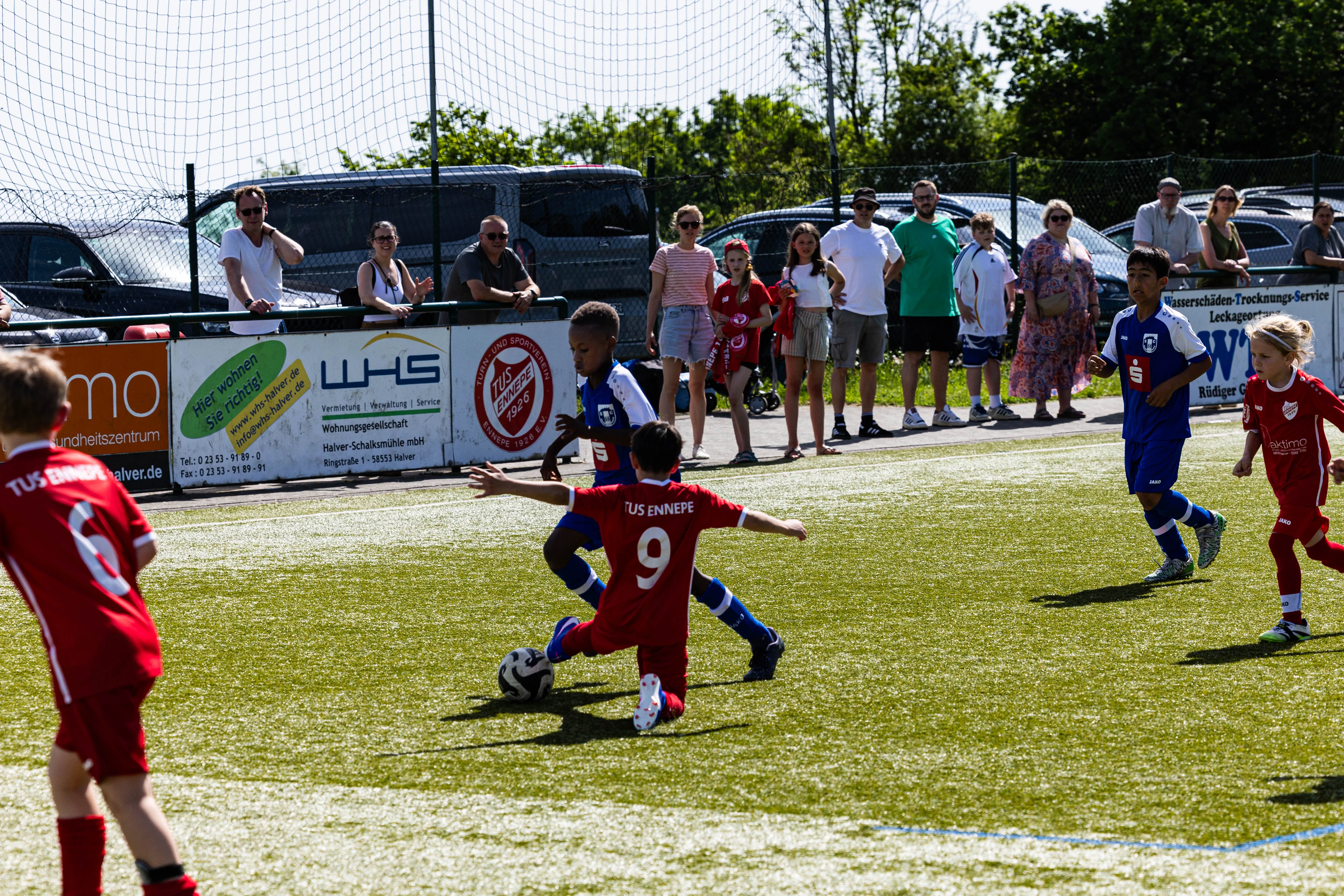 Zwei Fußballspieler in roten Trikots kämpfen um den Ball, während im Hintergrund Zuschauer das Spiel verfolgen.