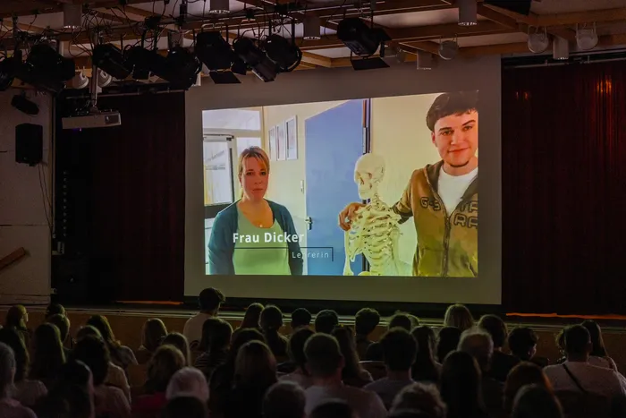 Ein Bild von einer Projektion auf einer Bühne zeigt eine Lehrerin und einen Schüler, die ein Skelett halten. Im Publikum sitzen viele Menschen.