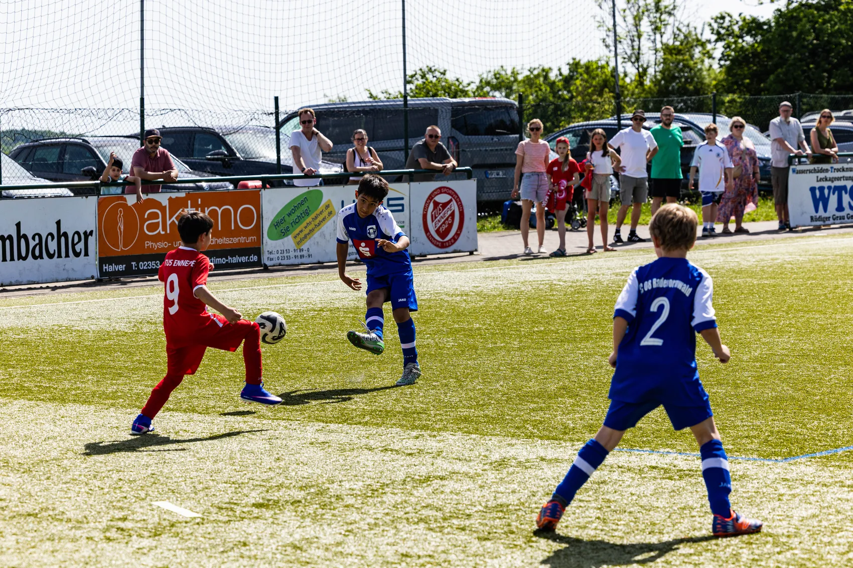 Zwei Fußballspieler in roten und blauen Trikots kämpfen um den Ball während eines Jugendfußballturniers, im Hintergrund stehen Zuschauer am Spielfeldrand.