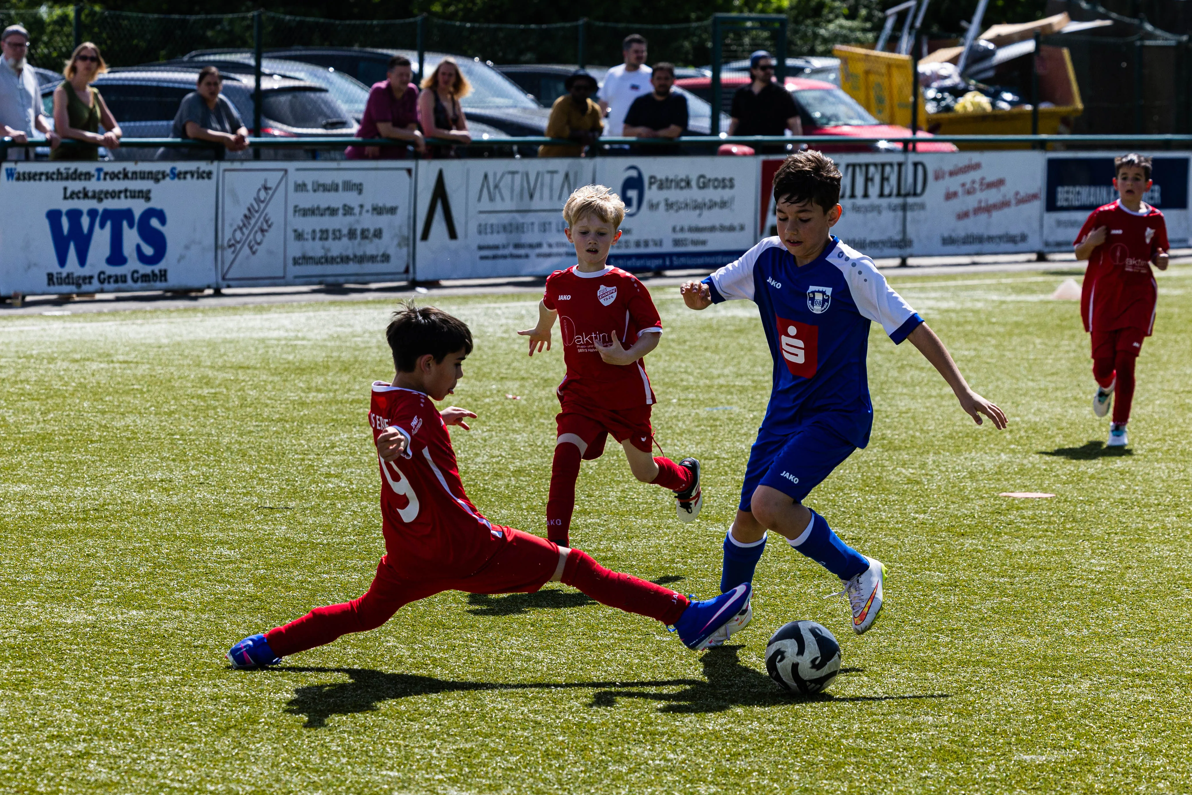 Zwei Jugendfußballspieler im Spiel: Ein Spieler in Rot versucht, den Ball von einem Spieler in Blau zu erobern. Im Hintergrund stehen Zuschauer und weitere Spieler auf dem Rasen.