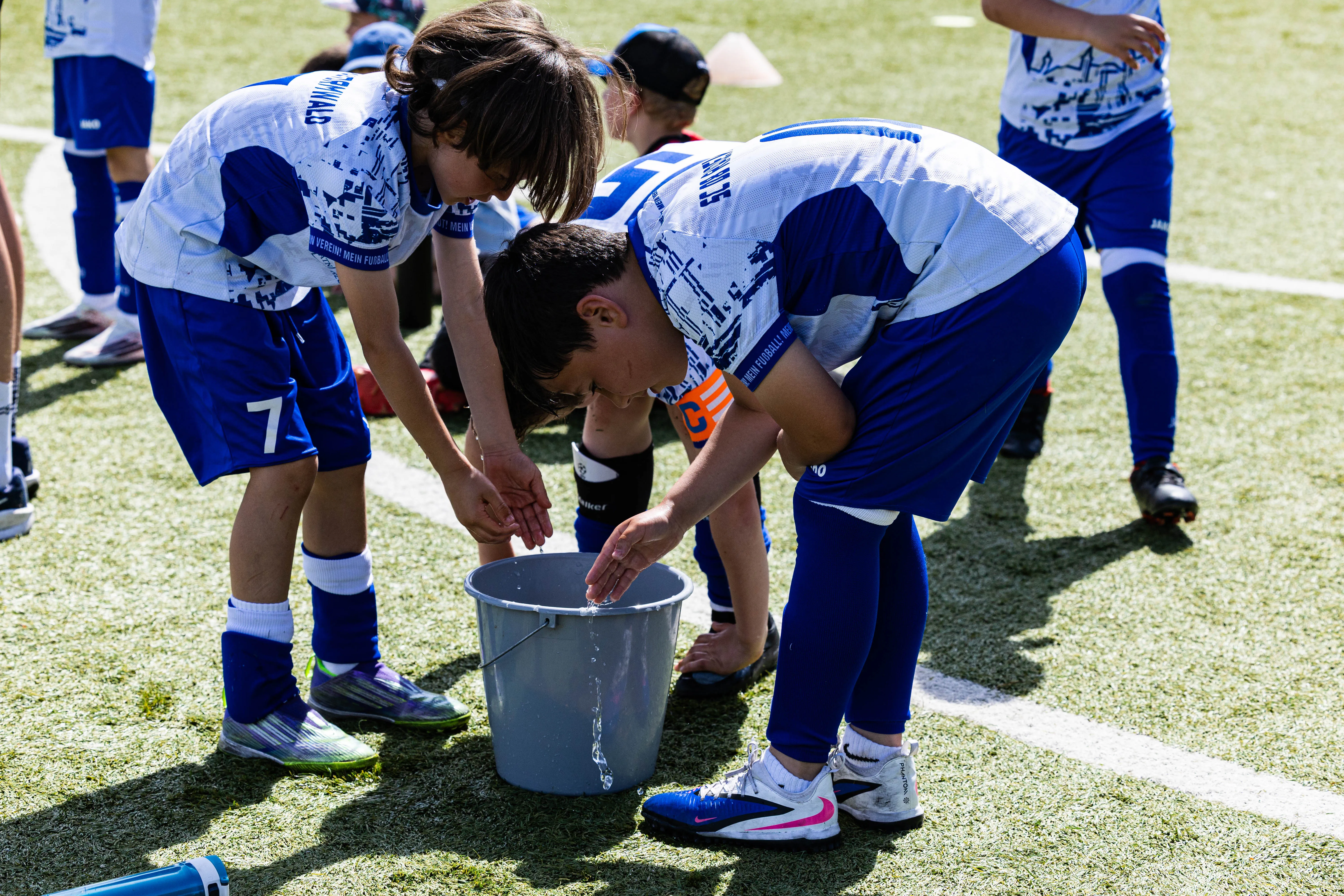 Drei junge Fußballspieler in blauen Trikots und Shorts beugen sich über einen grauen Eimer, vermutlich um Wasser zu holen.