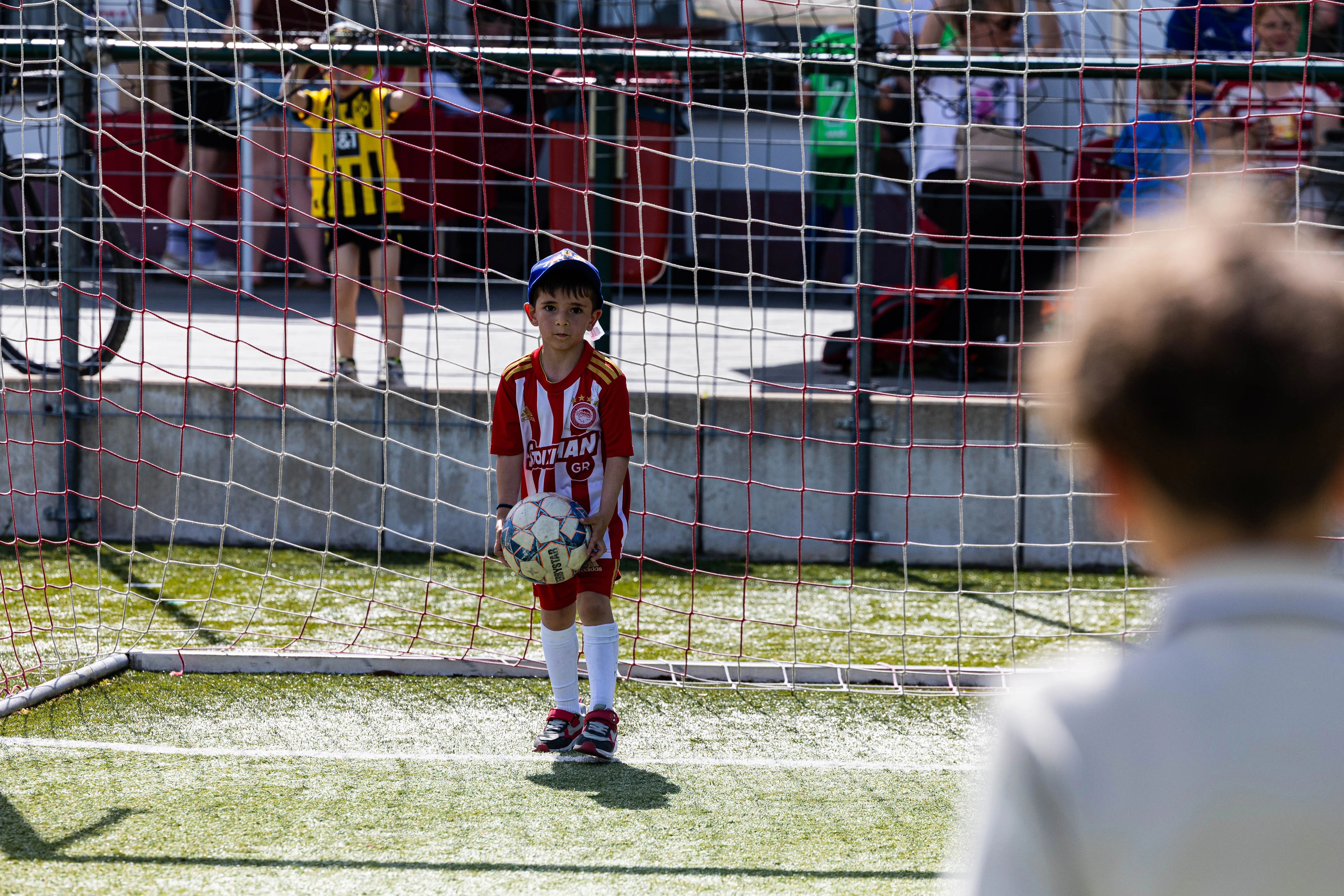 Junge im Torwarttrikot mit Ball steht auf einem Fußballfeld vor dem Tor. Im Hintergrund sind Zuschauer und weitere Spieler zu sehen.