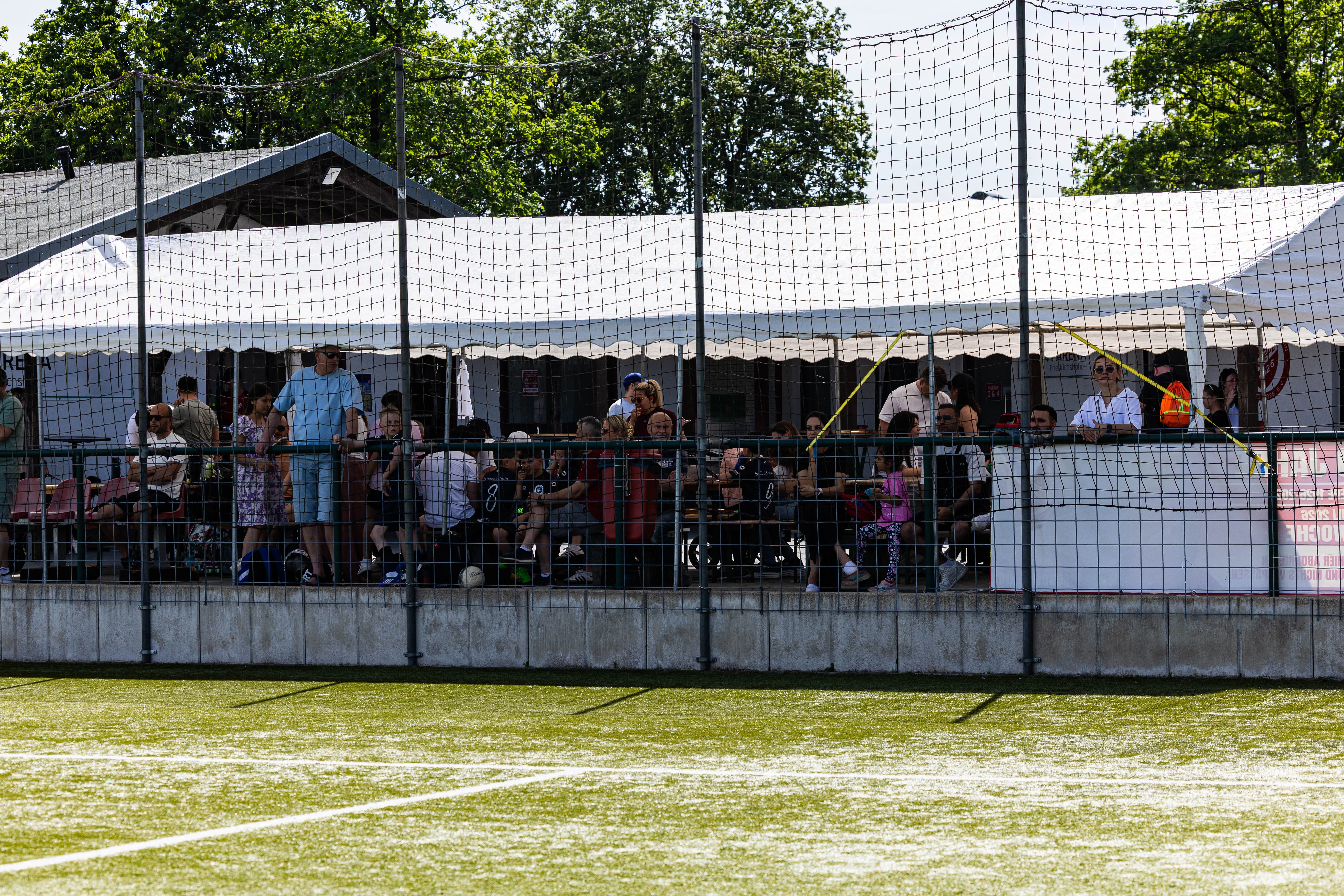 Zuschauer stehen hinter einem Maschendrahtzaun und beobachten ein Fußballspiel auf einem grünen Kunstrasenplatz.