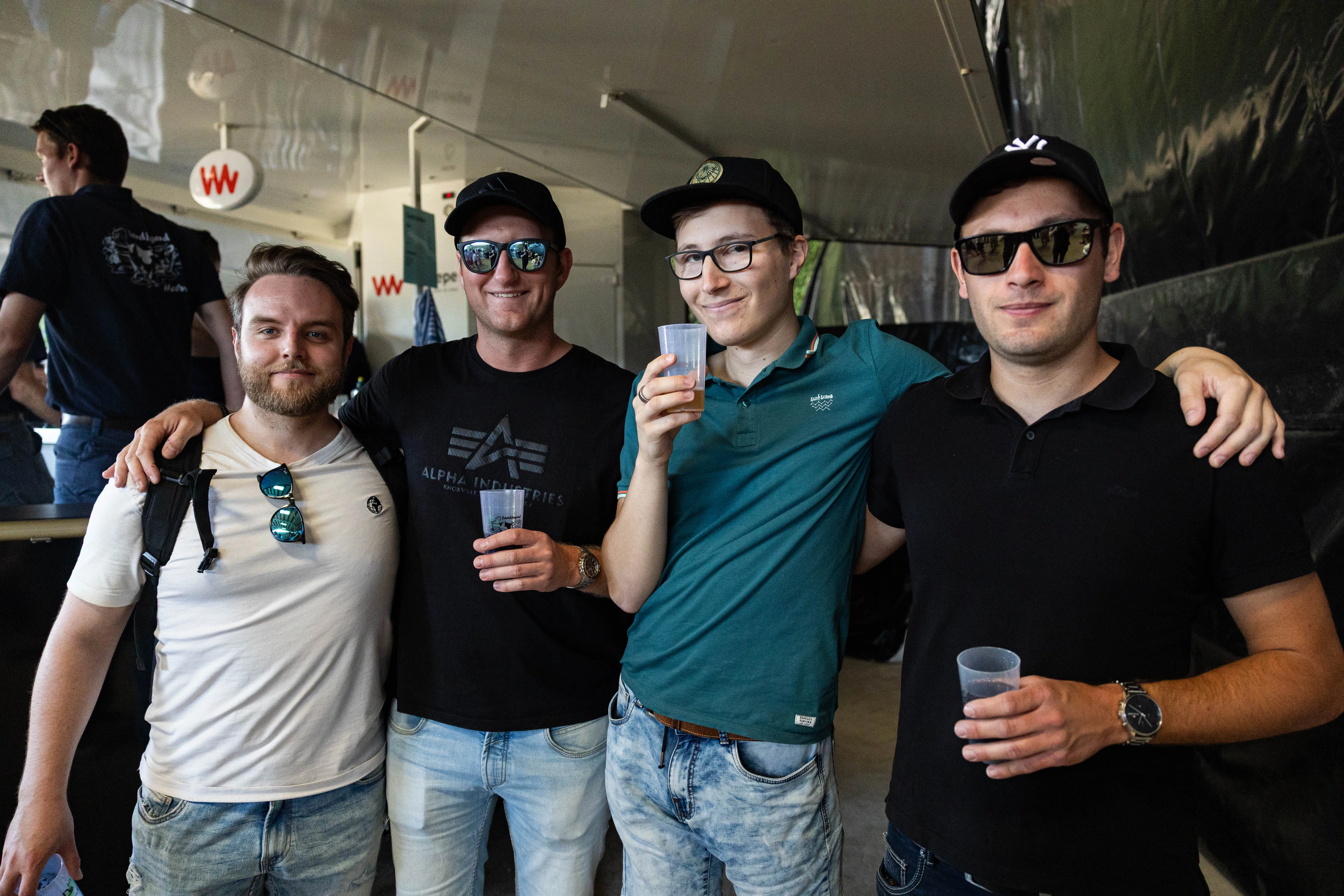 Vier junge Männer stehen eng zusammen und lächeln in die Kamera, jeder hält ein Glas in der Hand. Im Hintergrund sind weitere Personen und ein Zelt mit dem Logo eines Sponsors zu sehen.