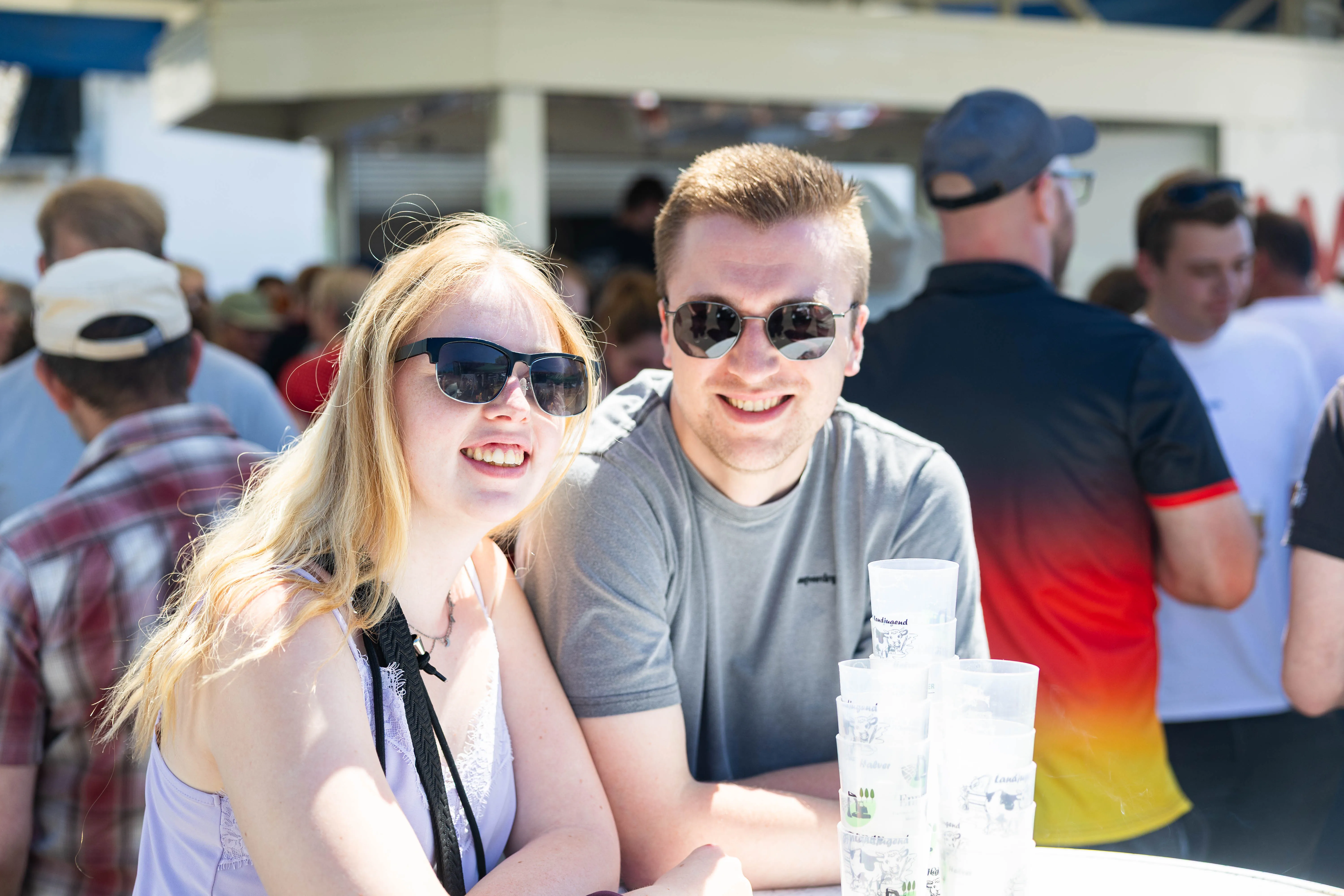 Zwei junge Erwachsene mit Sonnenbrillen lächeln in die Kamera, vor ihnen gestapelte Plastikbecher. Im Hintergrund sind weitere Menschen auf einem Hof zu sehen.