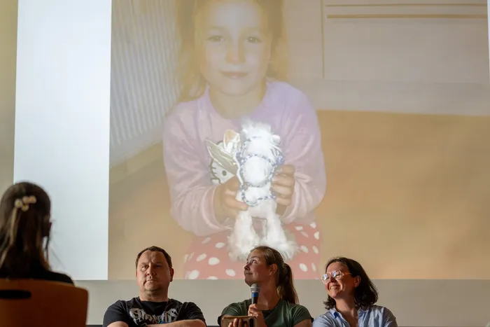 Ein Mädchen hält ein selbstgebasteltes Tier vor der Kamera. Darunter sitzen drei Erwachsene, von denen eine in die Kamera spricht. Im Hintergrund wird das Bild des Mädchens auf eine Leinwand projiziert.