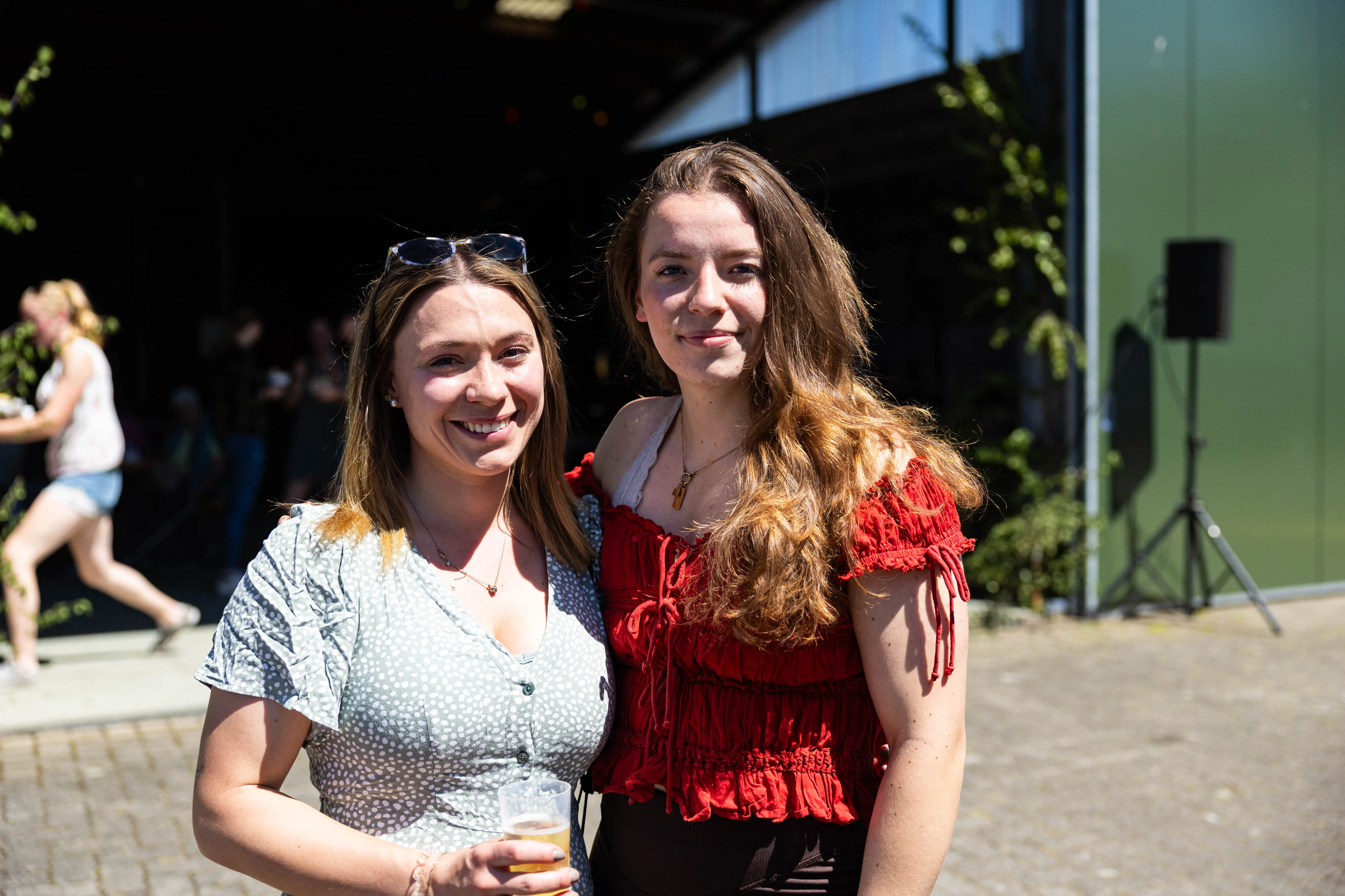 Zwei junge Frauen lächeln in die Kamera, jede hält ein Glas Bier. Im Hintergrund sind weitere Menschen und ein Gebäude zu sehen.