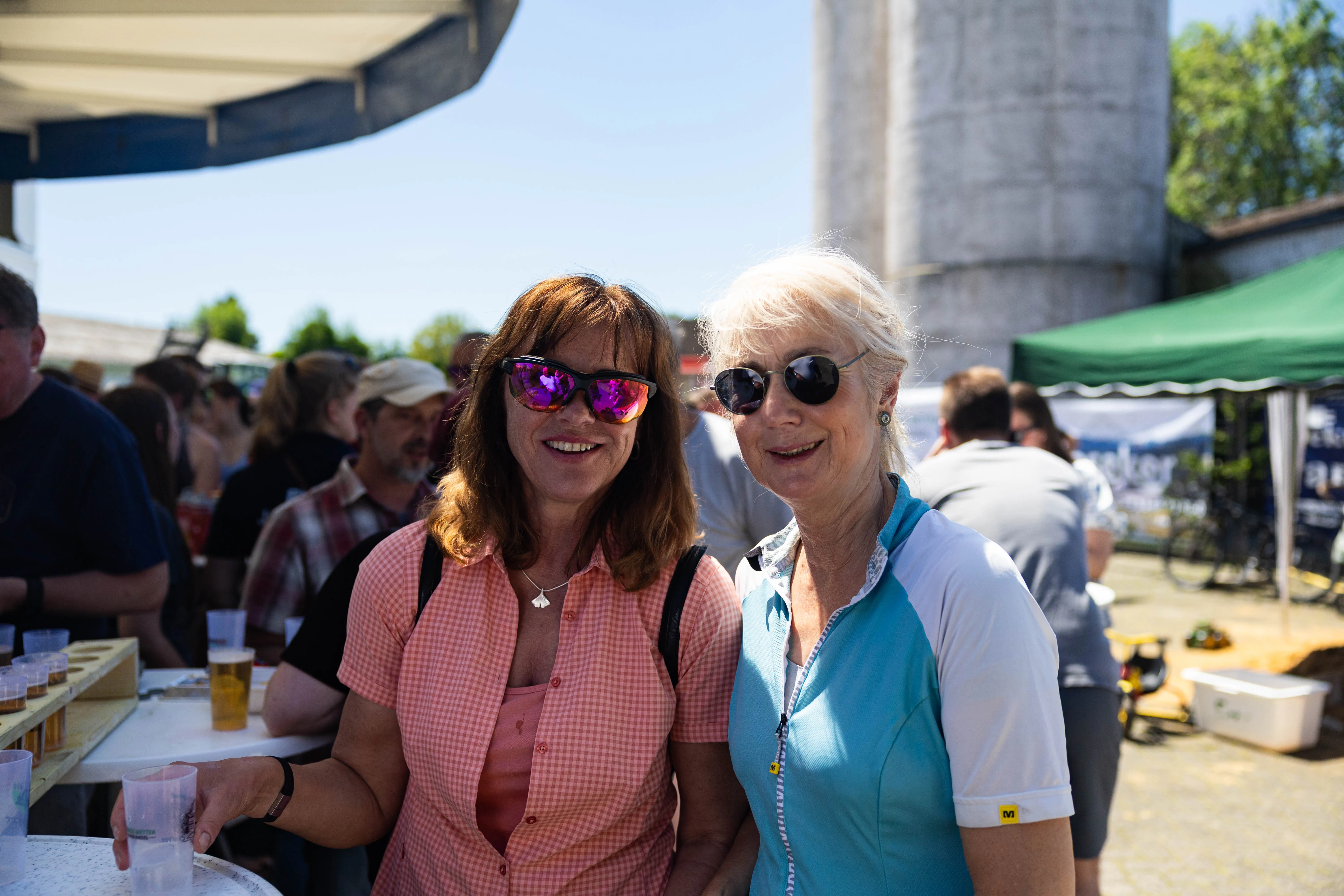 Zwei lächelnde Frauen mit Sonnenbrille stehen vor einer Menschenmenge und einem großen Siloturm. Im Hintergrund sind Zelte und Tische zu sehen.