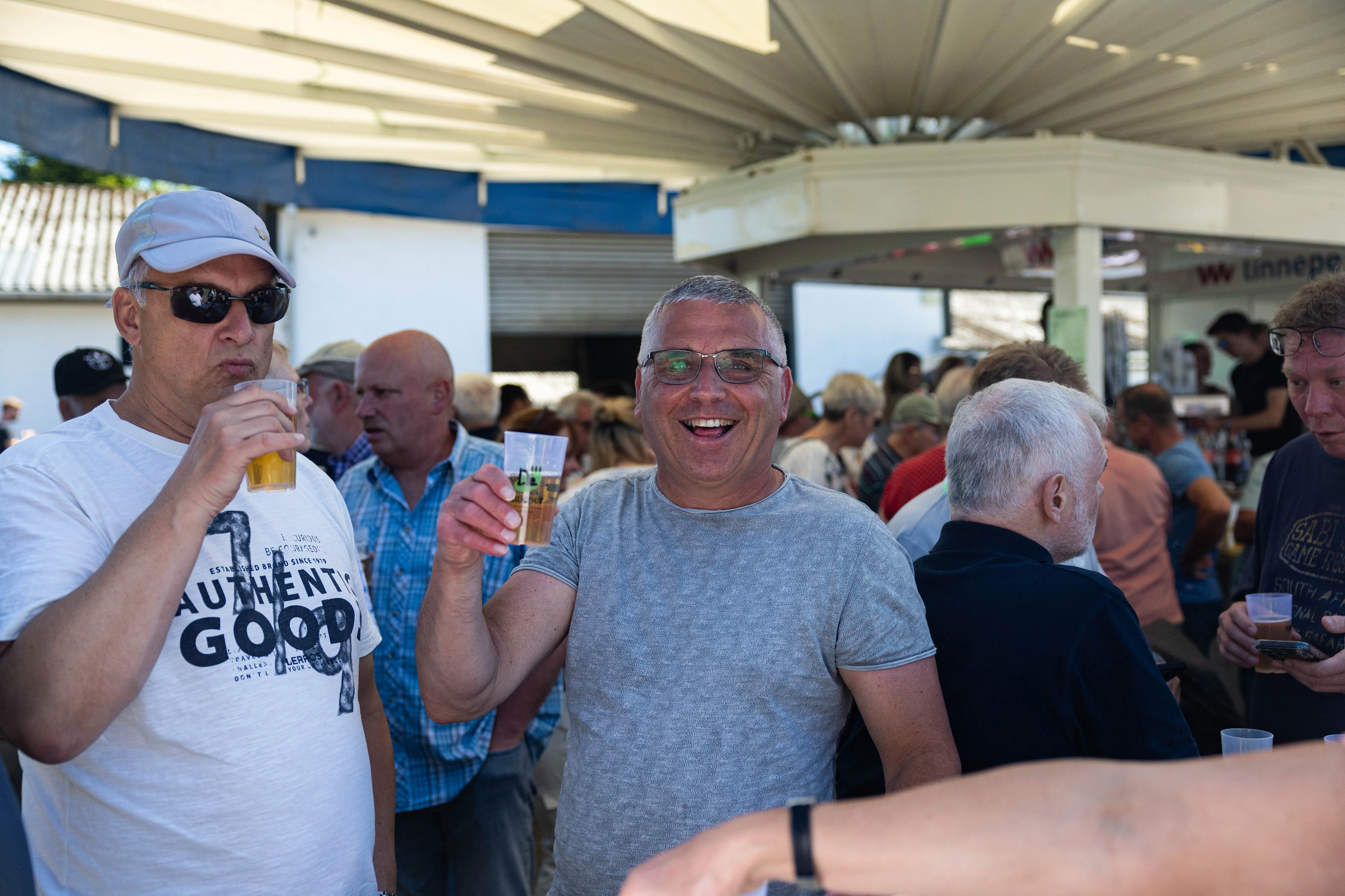 Zwei Männer lächeln in die Kamera, während sie auf einem Hof bei einem Frühschoppen Bier trinken. Im Hintergrund sind weitere Menschen und ein weißes Zelt zu sehen.