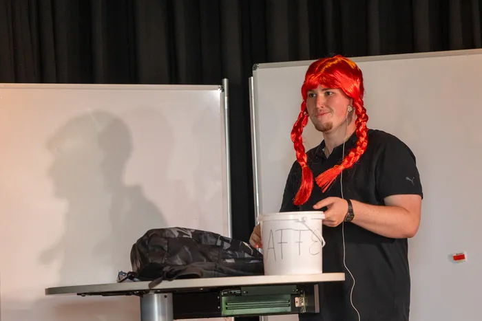 Ein junger Mann mit rotem Perückenhaar und zwei Zöpfen steht auf einer Bühne vor einer weißen Leinwand und hält einen weißen Eimer mit der Aufschrift 