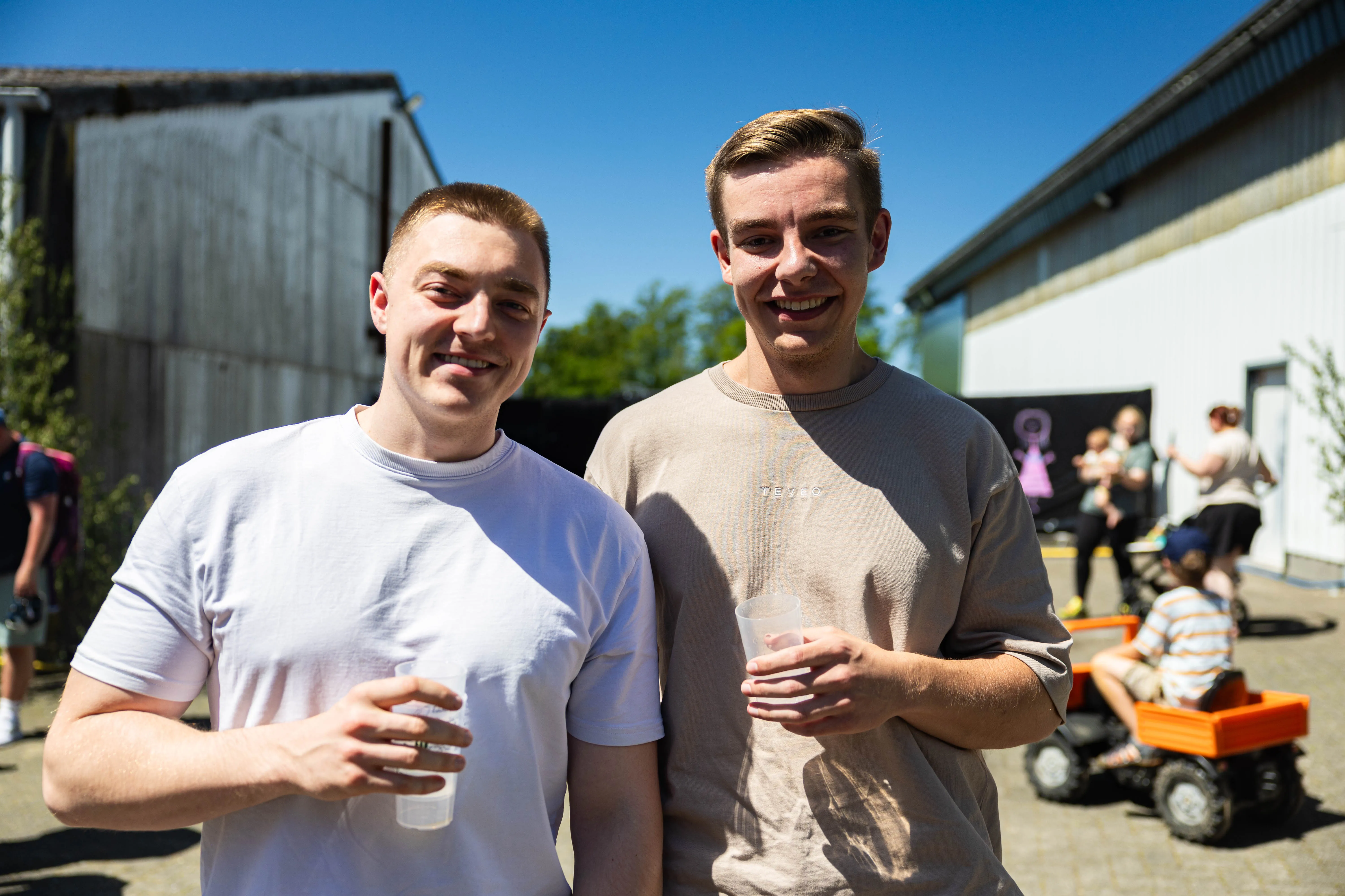 Zwei junge Männer lächeln in die Kamera, jeder hält ein Glas in der Hand. Im Hintergrund sind weitere Menschen und kleine Traktoren zu sehen.