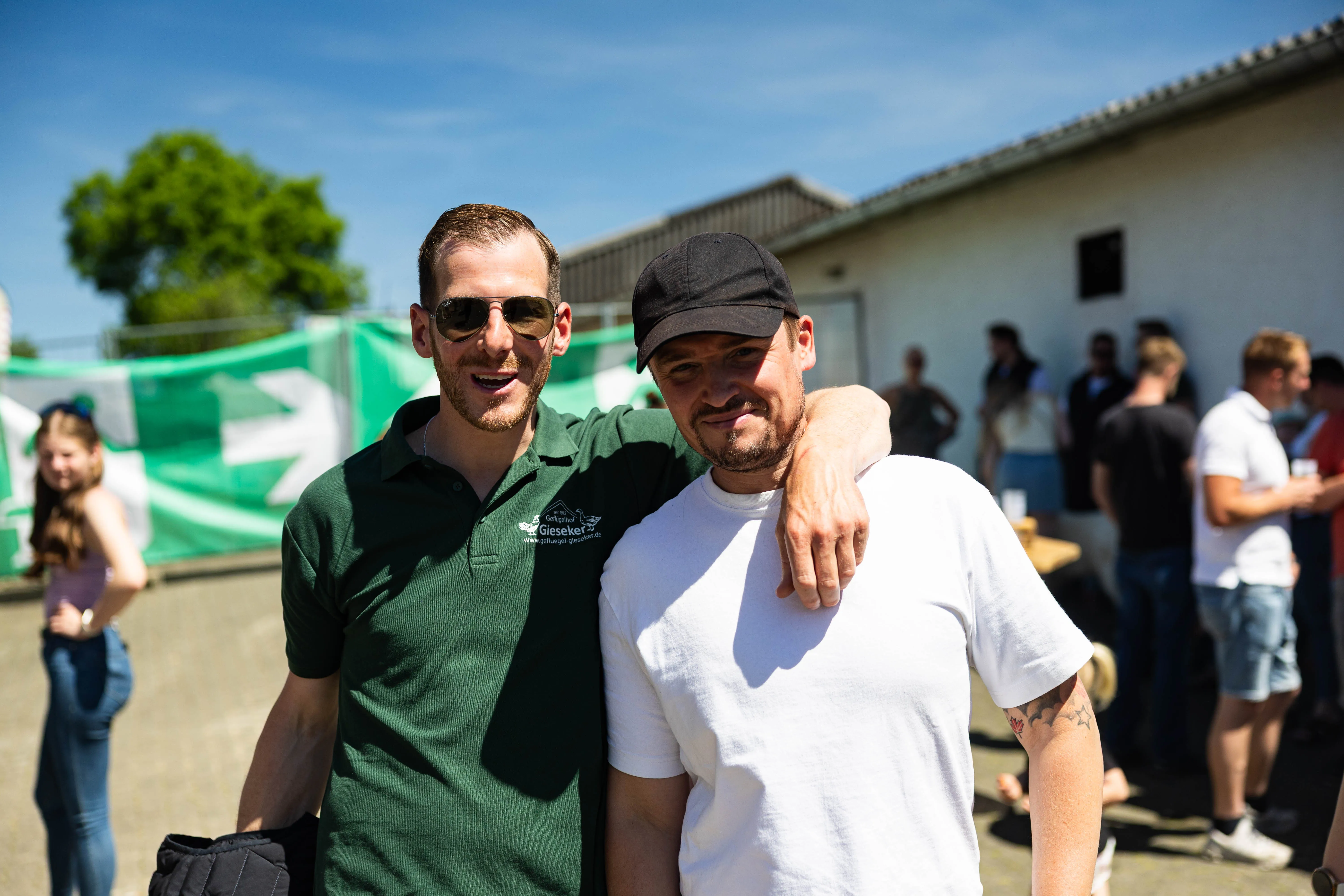 Zwei lächelnde Männer posieren eng nebeneinander, der Mann links trägt eine Sonnenbrille und ein grünes Poloshirt mit Logo, der andere eine Baseballkappe und ein weißes T-Shirt. Im Hintergrund sind weitere Menschen und Gebäude auf einem Hof zu sehen.