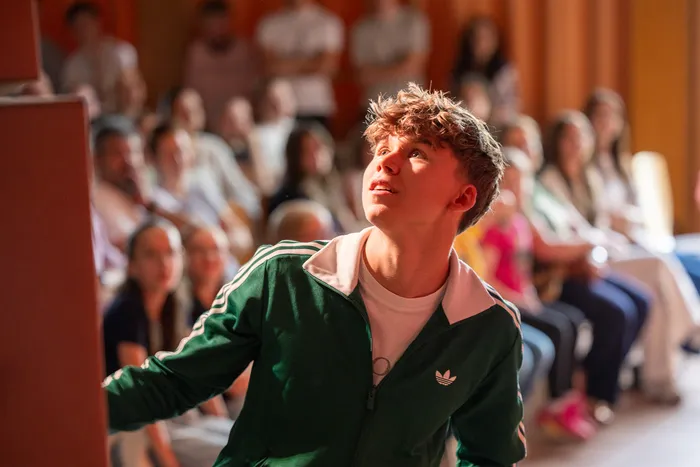 Ein junger Mann mit lockigem Haar blickt während einer Veranstaltung in einem Raum mit vielen Zuschauern nach oben. Er trägt eine grüne Jacke mit weißen Streifen.