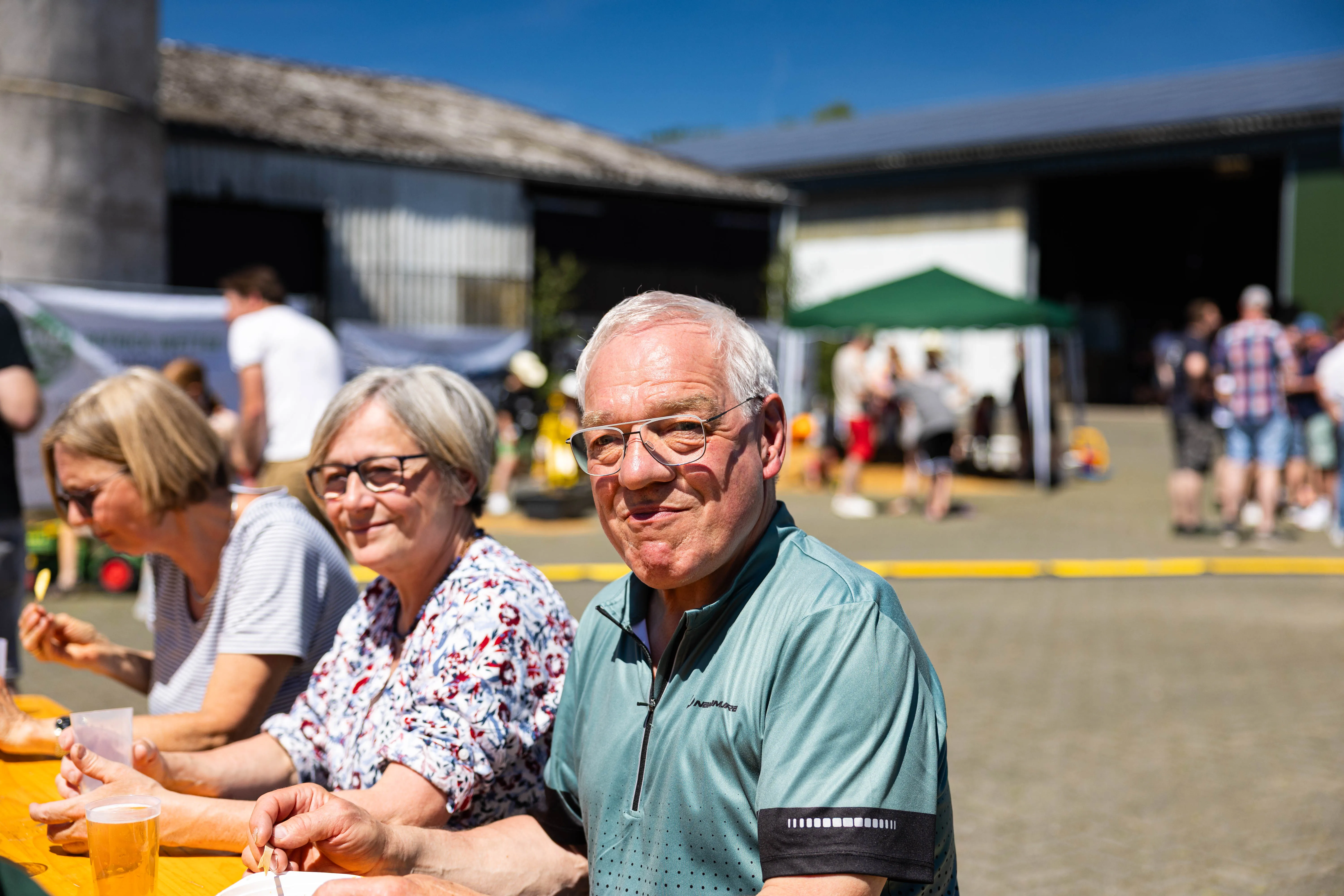 Ein älterer Mann mit Brille lächelt in die Kamera, während er mit anderen Personen an einem Biergartentisch sitzt. Im Hintergrund sind weitere Besucher und Gebäude eines Bauernhofs zu sehen.