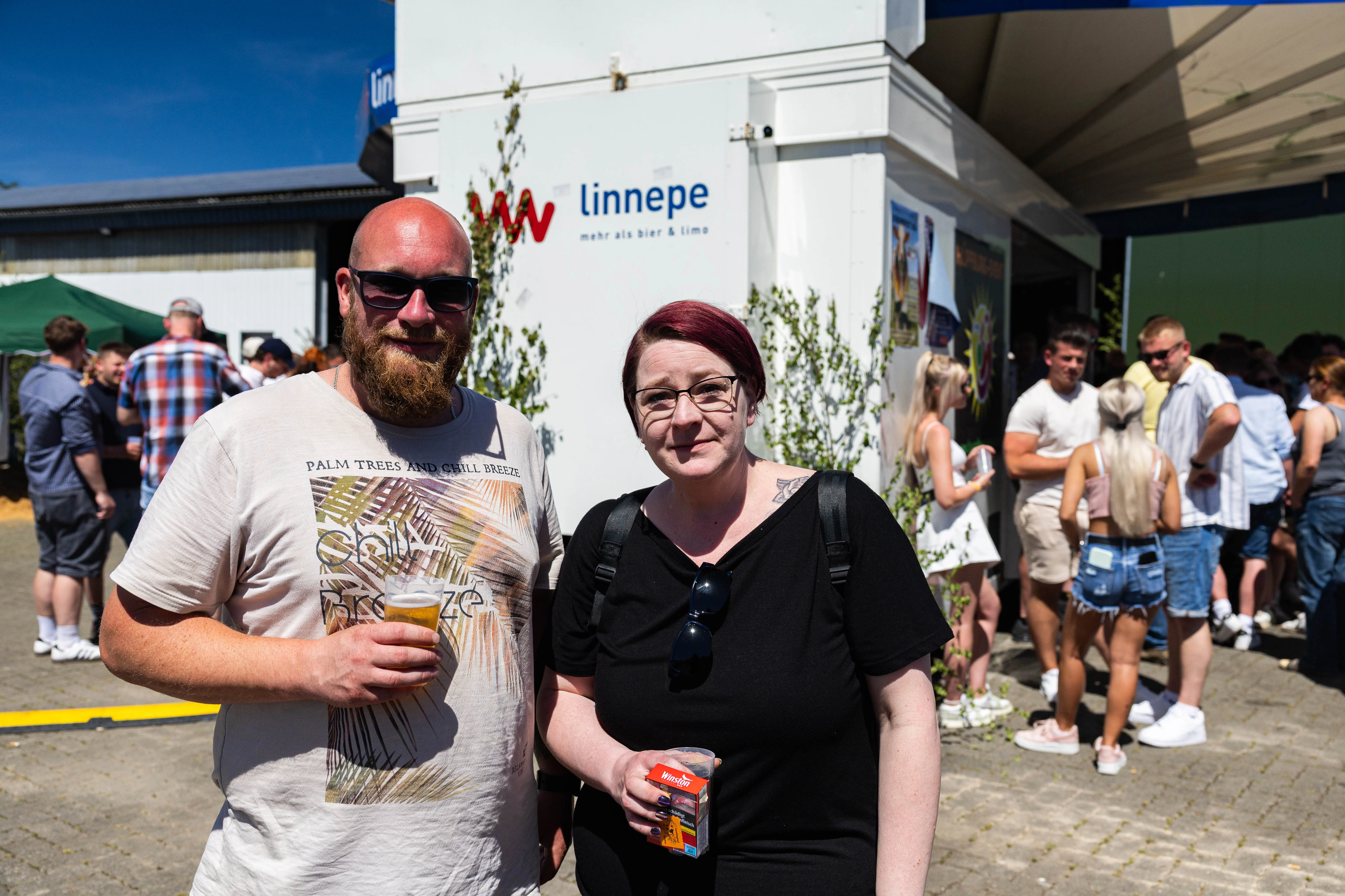 Ein Mann mit Bart und eine Frau mit roten Haaren stehen lächelnd vor der Kamera und halten jeweils ein Glas Bier. Im Hintergrund sind weitere Menschen und ein Getränkestand zu sehen.