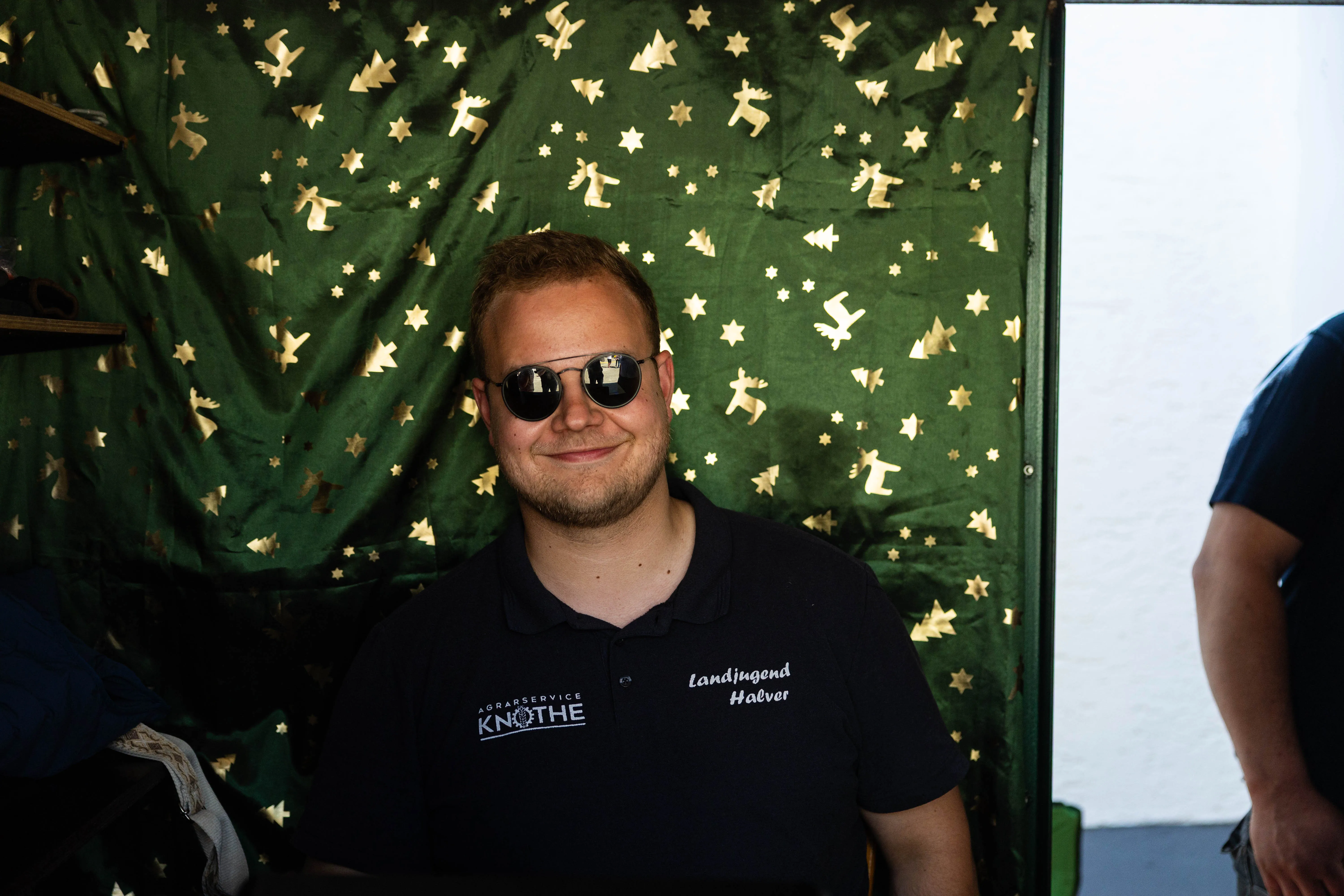 Ein junger Mann mit dunkler Sonnenbrille und Polohemd mit Aufschrift „Landjugend Halver“ lächelt in die Kamera. Im Hintergrund eine Wand mit Sternenmuster.