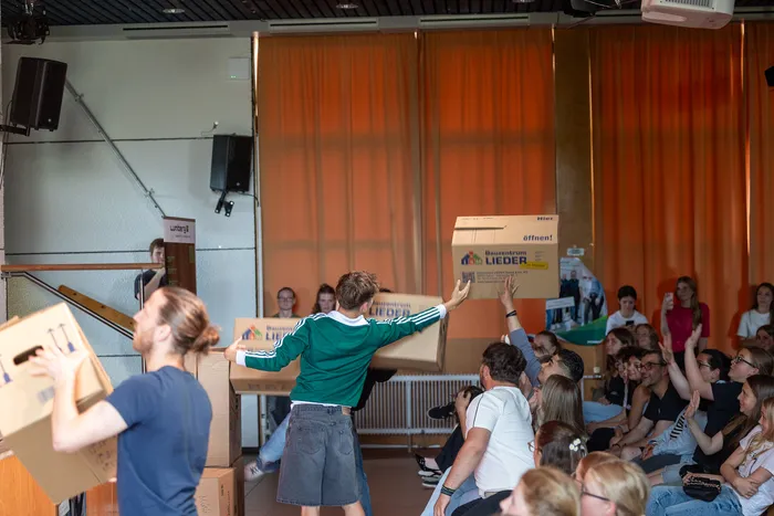 Ein junger Mann in Shorts und grünem Pullover steht auf einer Ansammlung von Kartons und hält einen weiteren Karton hoch. Im Hintergrund sitzen und stehen zahlreiche Zuschauer in einem großen Raum.