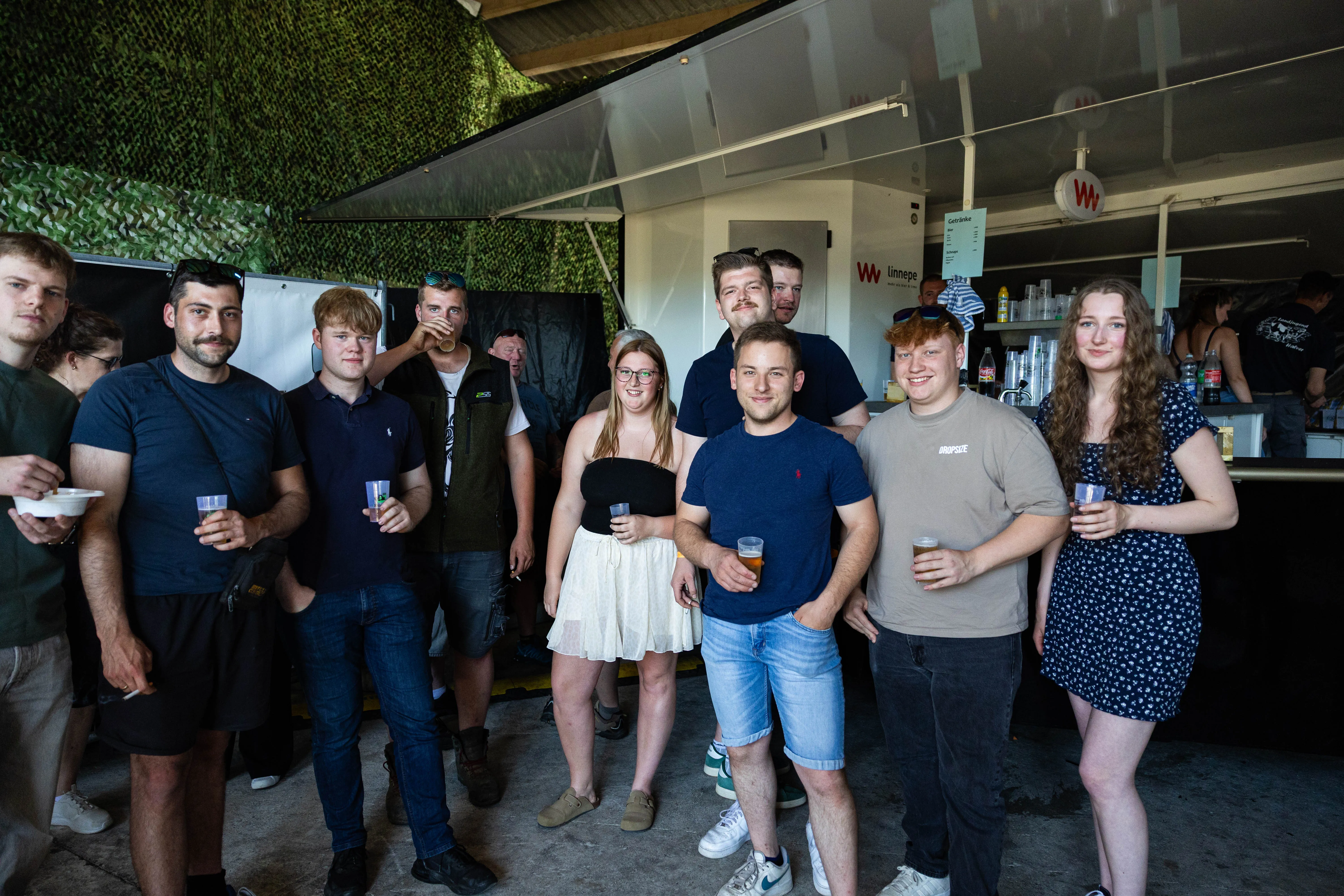 Eine Gruppe junger Erwachsener steht lächelnd mit Biergläsern vor einer provisorischen Bar im Freien. Im Hintergrund ist eine grüne Wand und ein Gebäude mit Werbeaufdruck zu sehen.