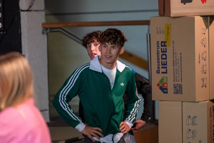 Ein junger Mann mit lockigem Haar trägt einen grünen Trainingsanzug und lächelt in die Kamera. Im Hintergrund sind Kartons mit der Aufschrift 