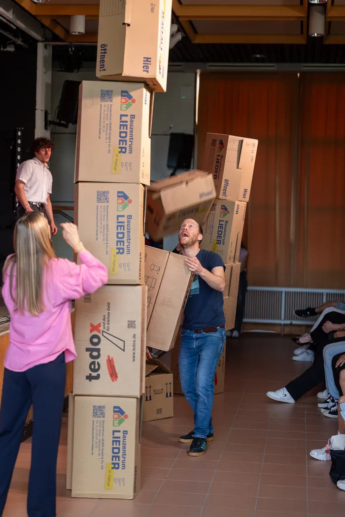 Ein Mann trägt einen hohen Turm aus Kartons mit der Aufschrift „Bautzentrum Lieder“ und „tedox“ auf seinem Kopf, während andere Menschen im Hintergrund zusehen. Der Raum scheint eine Turnhalle zu sein.