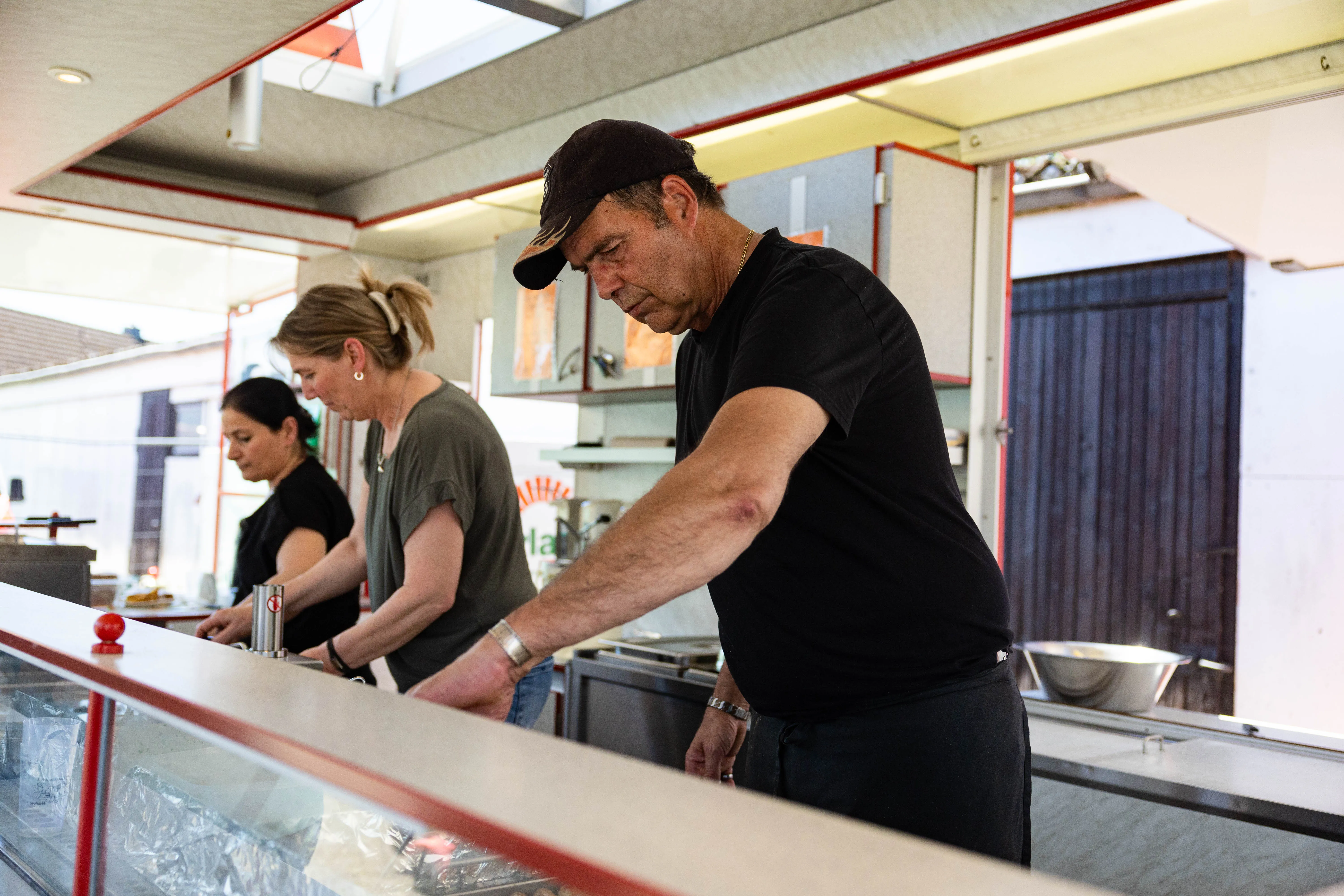 Zwei Personen arbeiten hinter einem Tresen, vermutlich in einer Imbissbude oder ähnlichem. Im Hintergrund sind weitere Personen und ein Blick in einen Raum erkennbar.