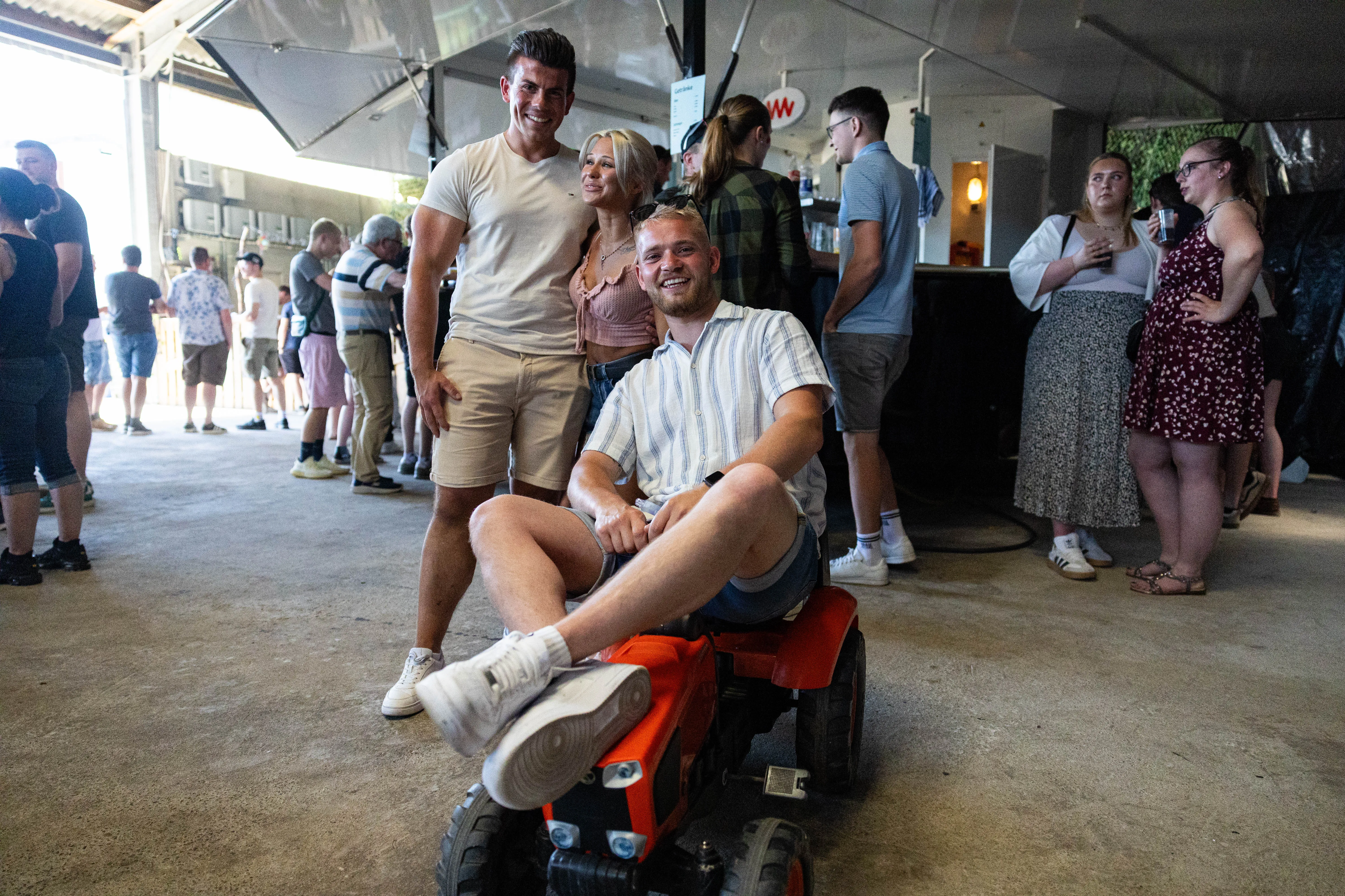Ein junger Mann sitzt lächelnd auf einem kleinen orangefarbenen Traktor vor einer Gruppe von Menschen, die an einer Bar stehen. Im Hintergrund ist eine offene Halle mit weiteren Besuchern zu sehen.