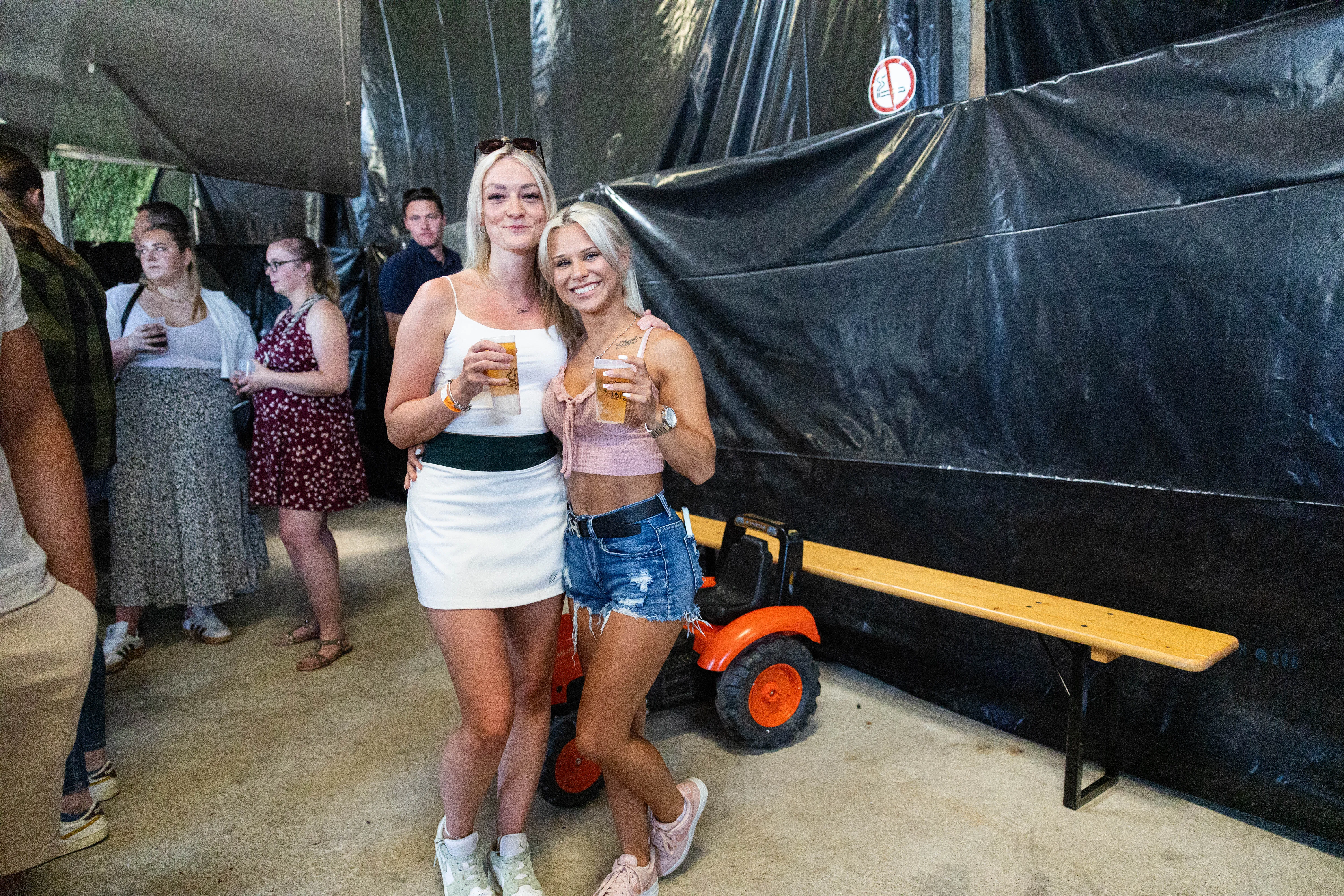 Zwei junge Frauen mit blonden Haaren stehen lächelnd vor einer dunklen Plane, jede hält ein Glas in der Hand. Im Hintergrund sind weitere Menschen und ein kleiner Traktor zu sehen.