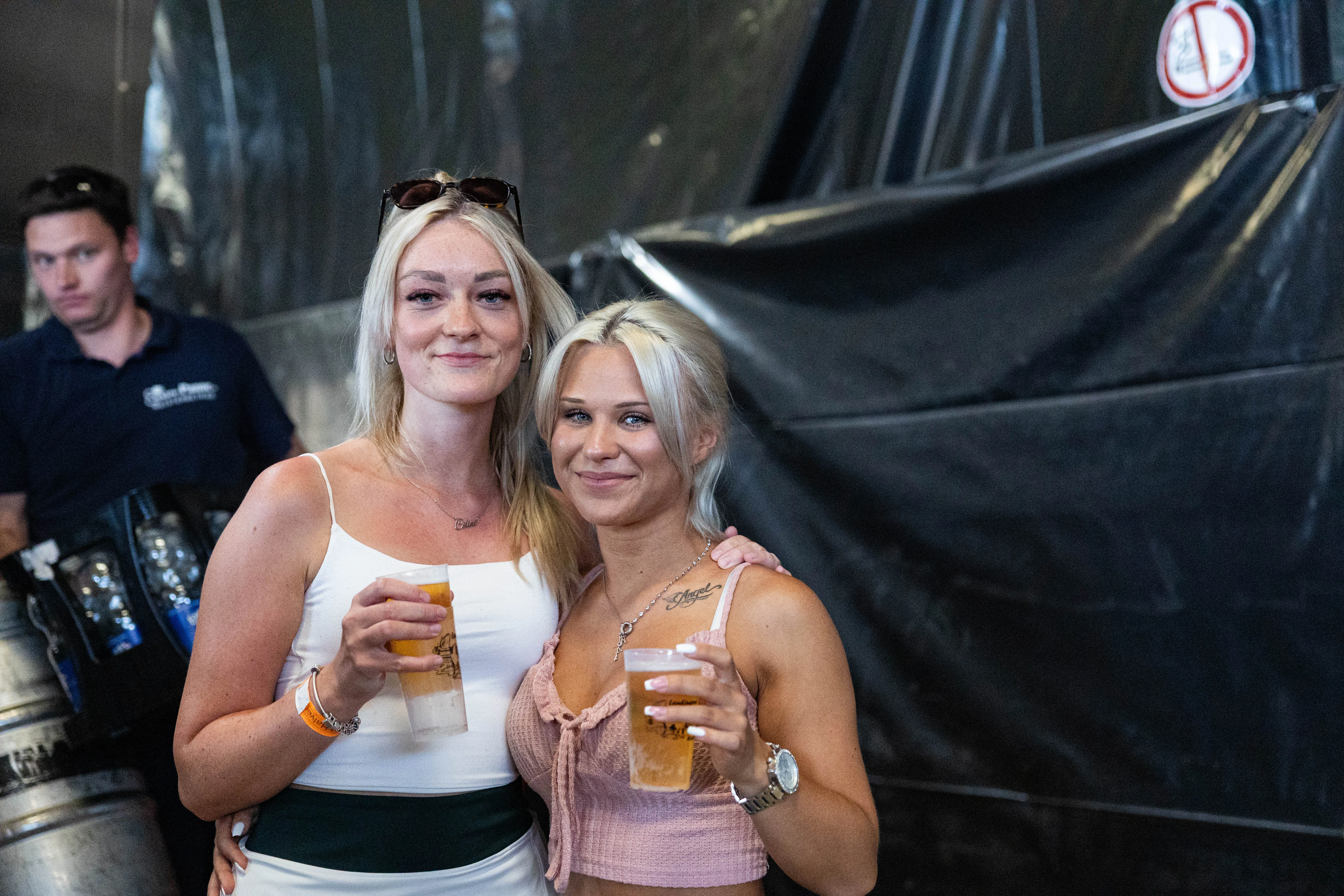 Zwei junge Frauen lächeln in die Kamera und halten jeweils ein Glas Bier in der Hand. Im Hintergrund steht ein Mann an einem Zapfhahn und ein dunkler Hintergrund.