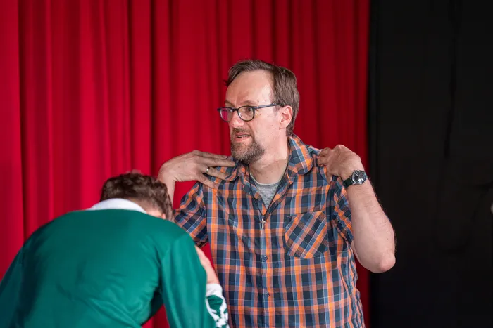 Ein Mann mit Bart und Brille in einem karierten Hemd wird von einer Person mit grünem Jackett an der Schulter berührt, während er spricht. Im Hintergrund ein roter Vorhang.